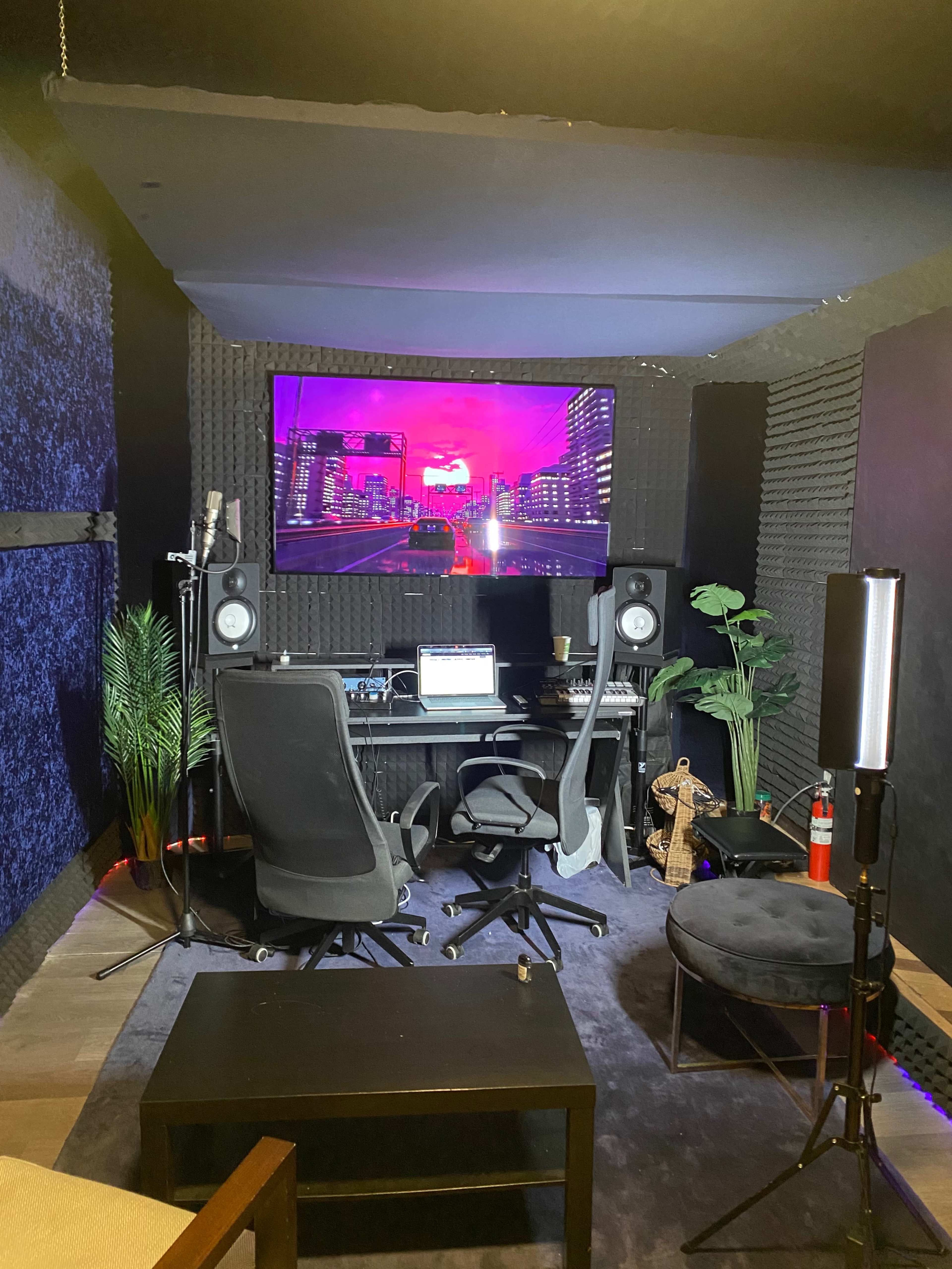 A recording studio with a desk set up for music production, featuring monitors, chairs, and acoustic foam panels on the walls.