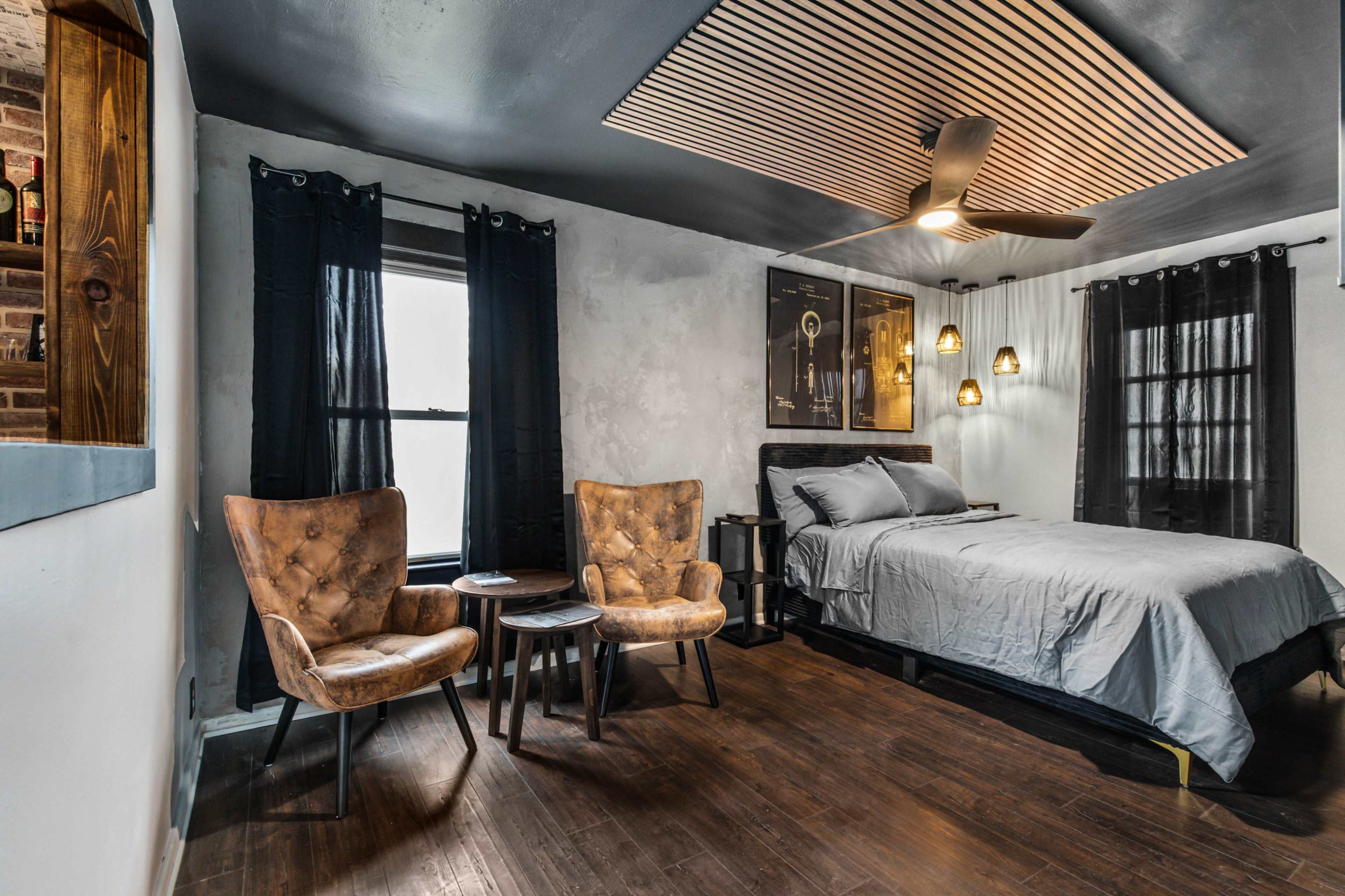 A bedroom featuring a bed, two armchairs, and a wooden ceiling design, with dark curtains and a wooden floor.
