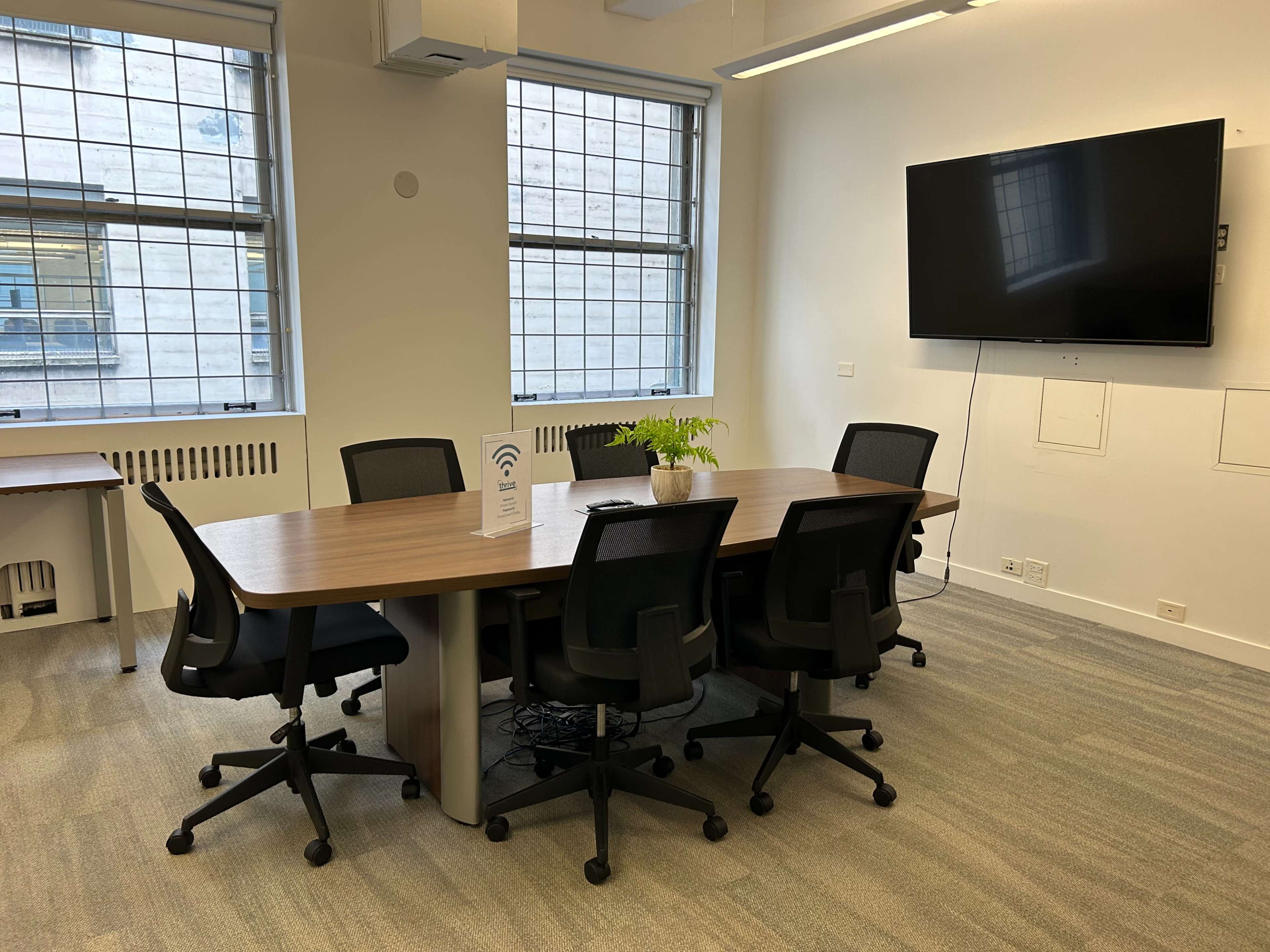 A conference room features a long table surrounded by six black rolling chairs, with a large screen mounted on the wall and windows allowing natural light.