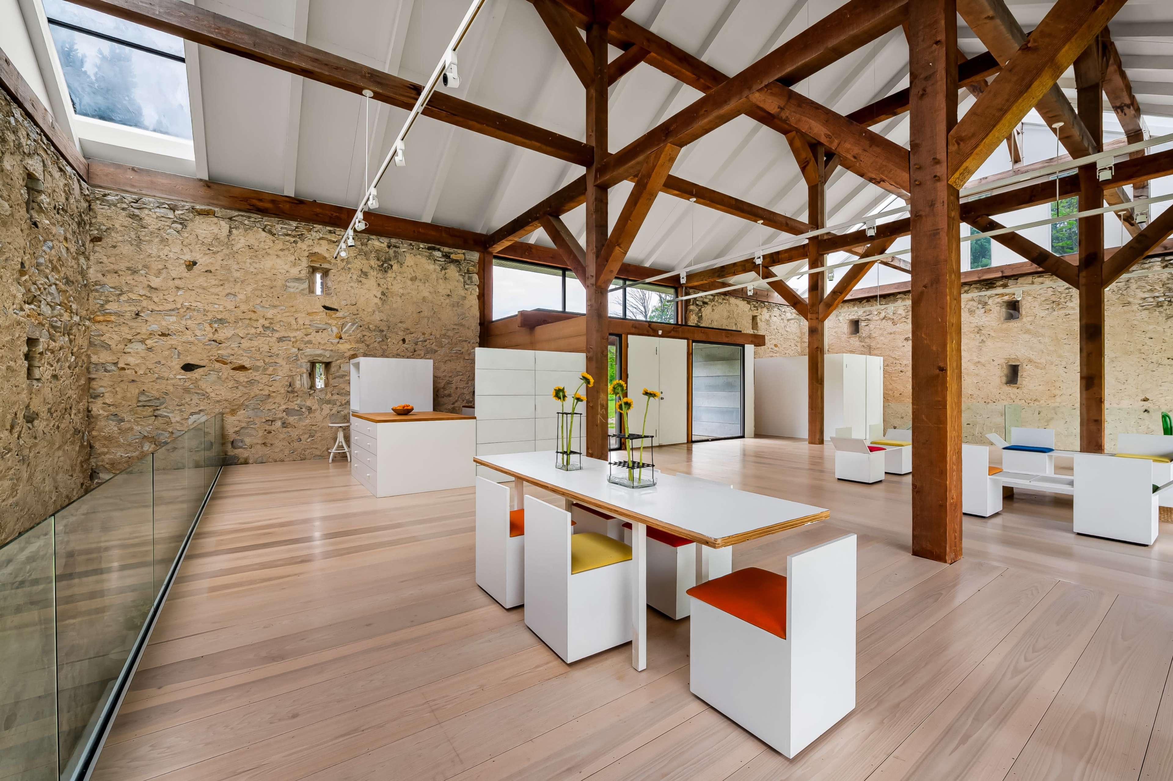 The image shows a modern interior space featuring wooden beams, stone walls, a large dining table with colorful chairs, and multiple glass panels.