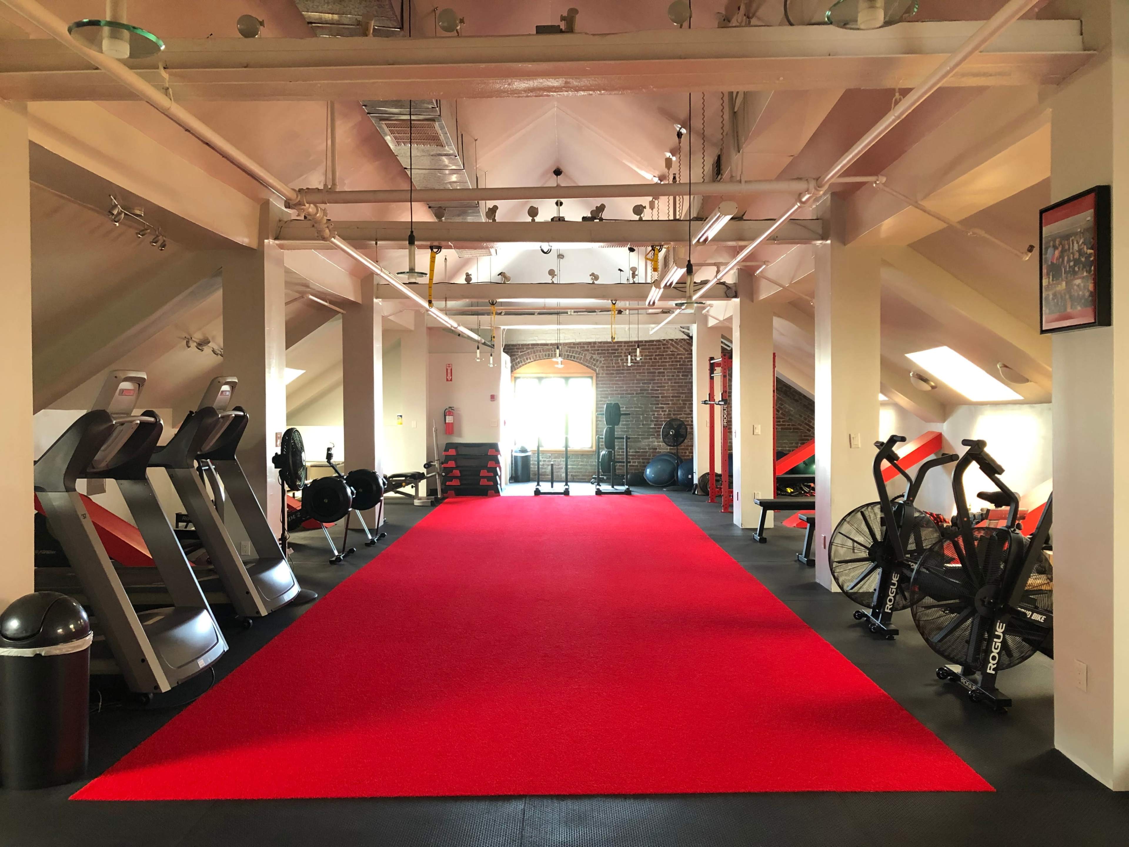 A well-lit gym with red carpet flooring, featuring several exercise machines, free weights, and workout equipment arranged along the walls.