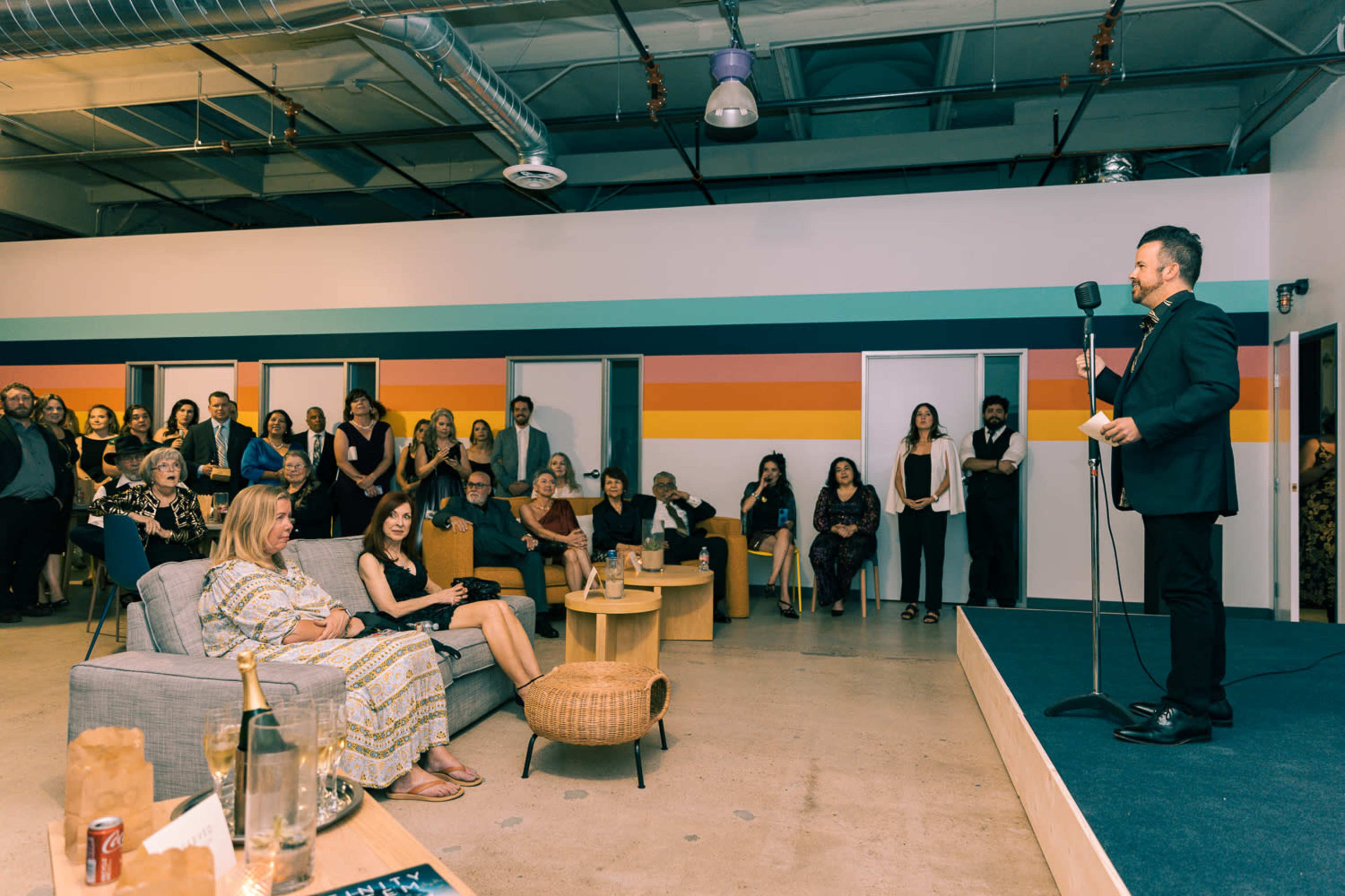 A man in a suit stands at a microphone while an audience sits in a brightly colored room with patterned walls.