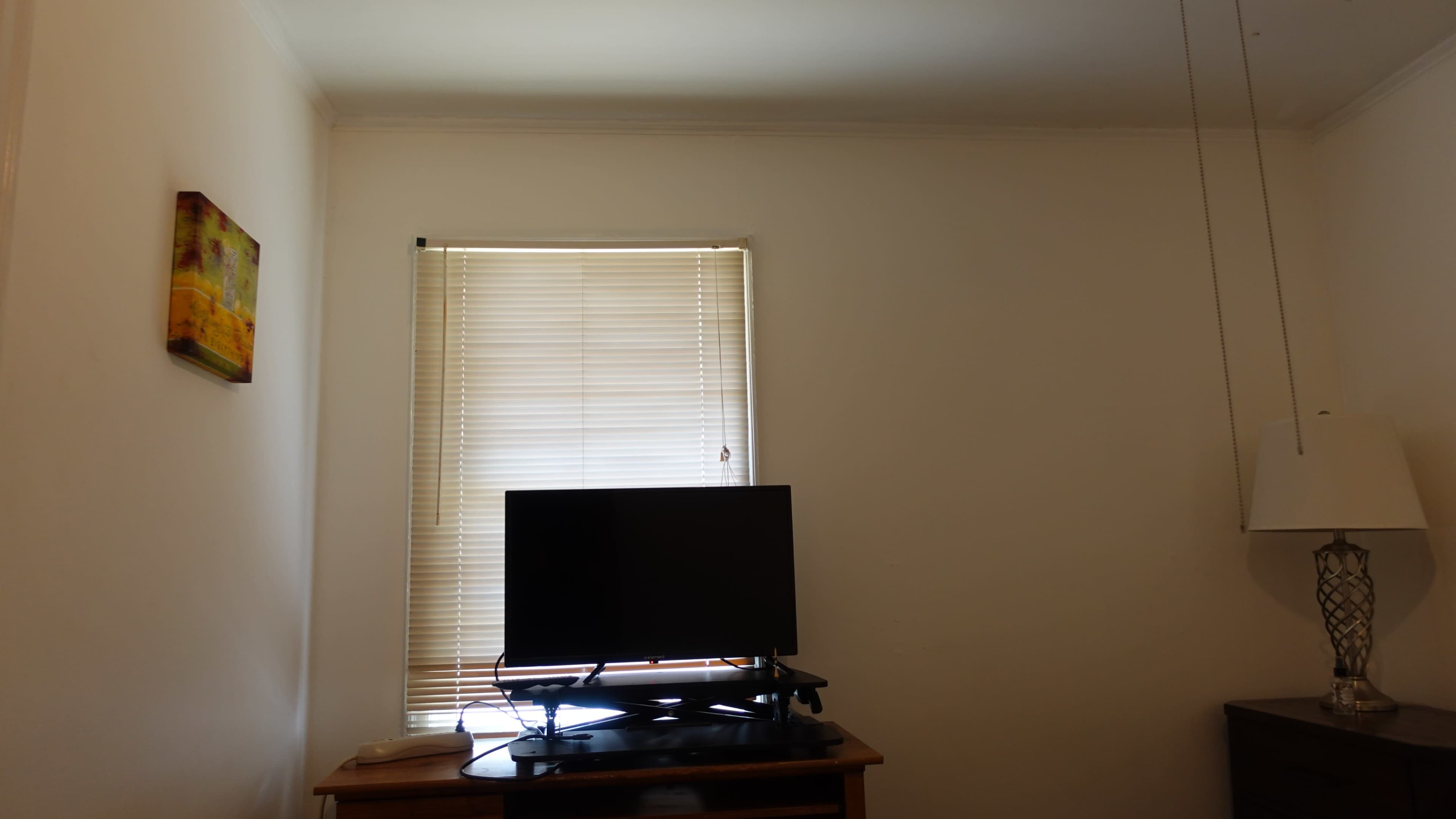 The image shows a room with a window covered by blinds, a television on a wooden stand, and a lamp beside it.
