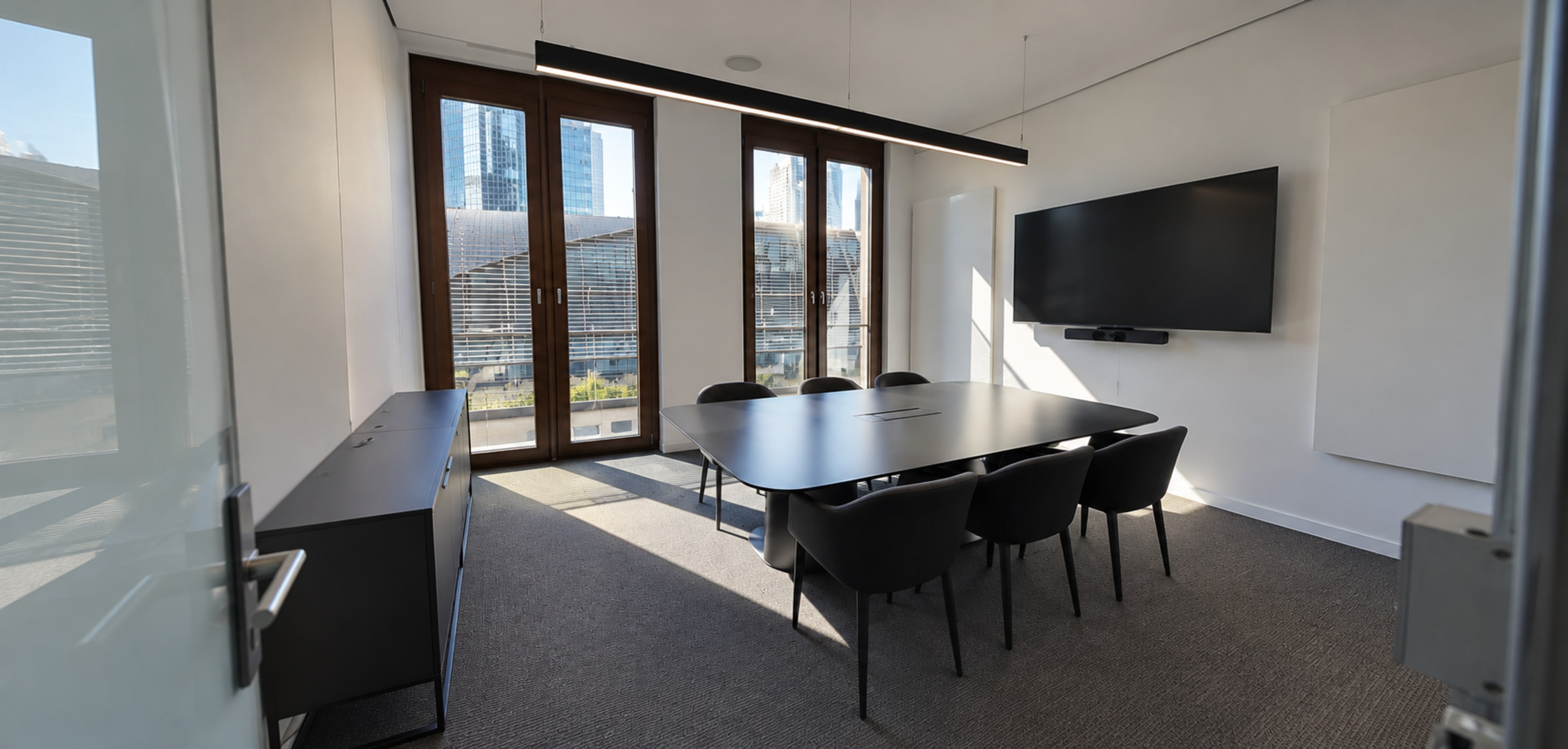 The image shows a modern conference room with a large table, six chairs, and a wall-mounted flat-screen display, featuring floor-to-ceiling windows that provide natural light.