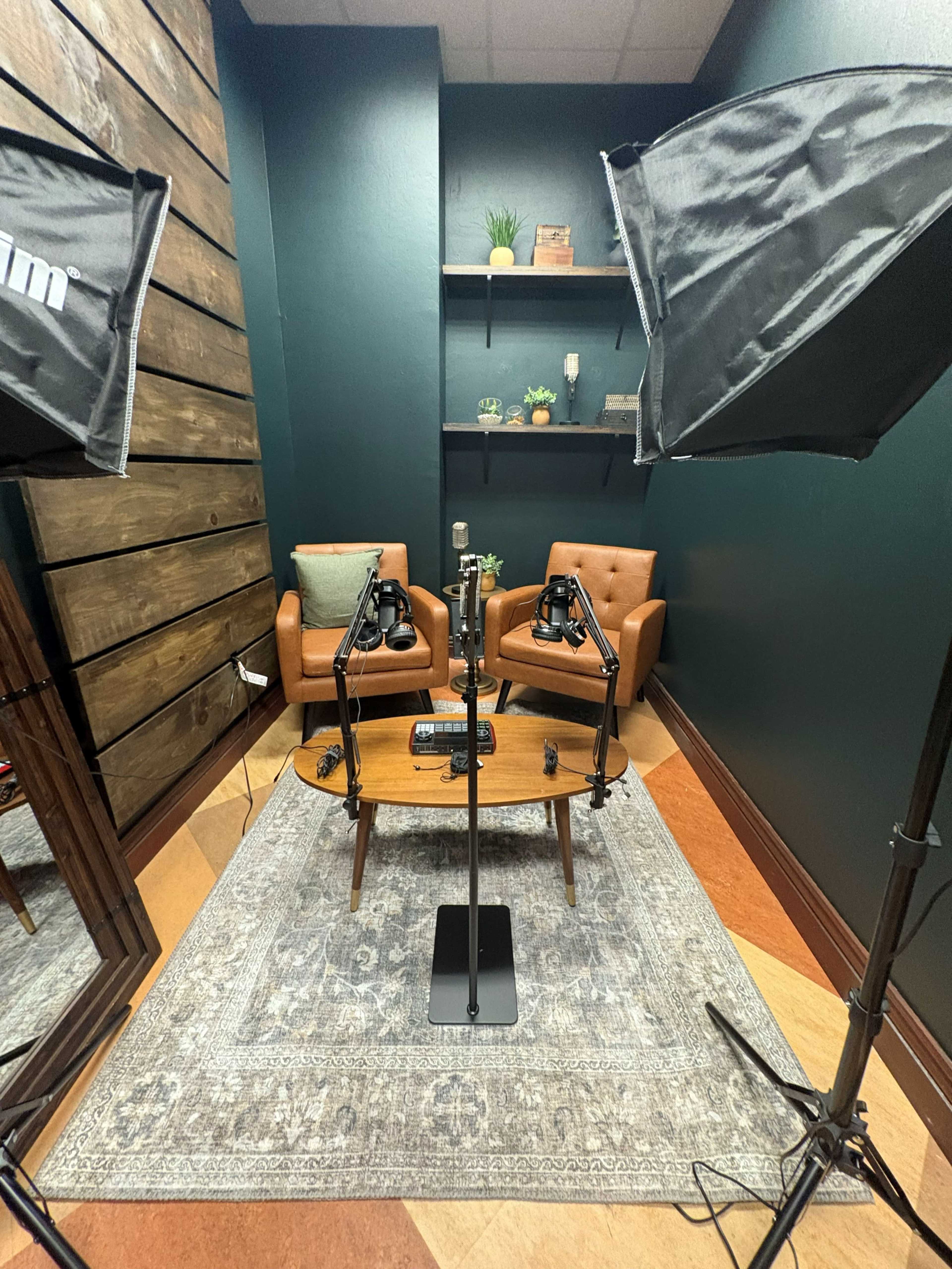 The image shows a small recording studio setup featuring two microphones on stands, a wooden table, two leather chairs, and two softbox lights positioned nearby.