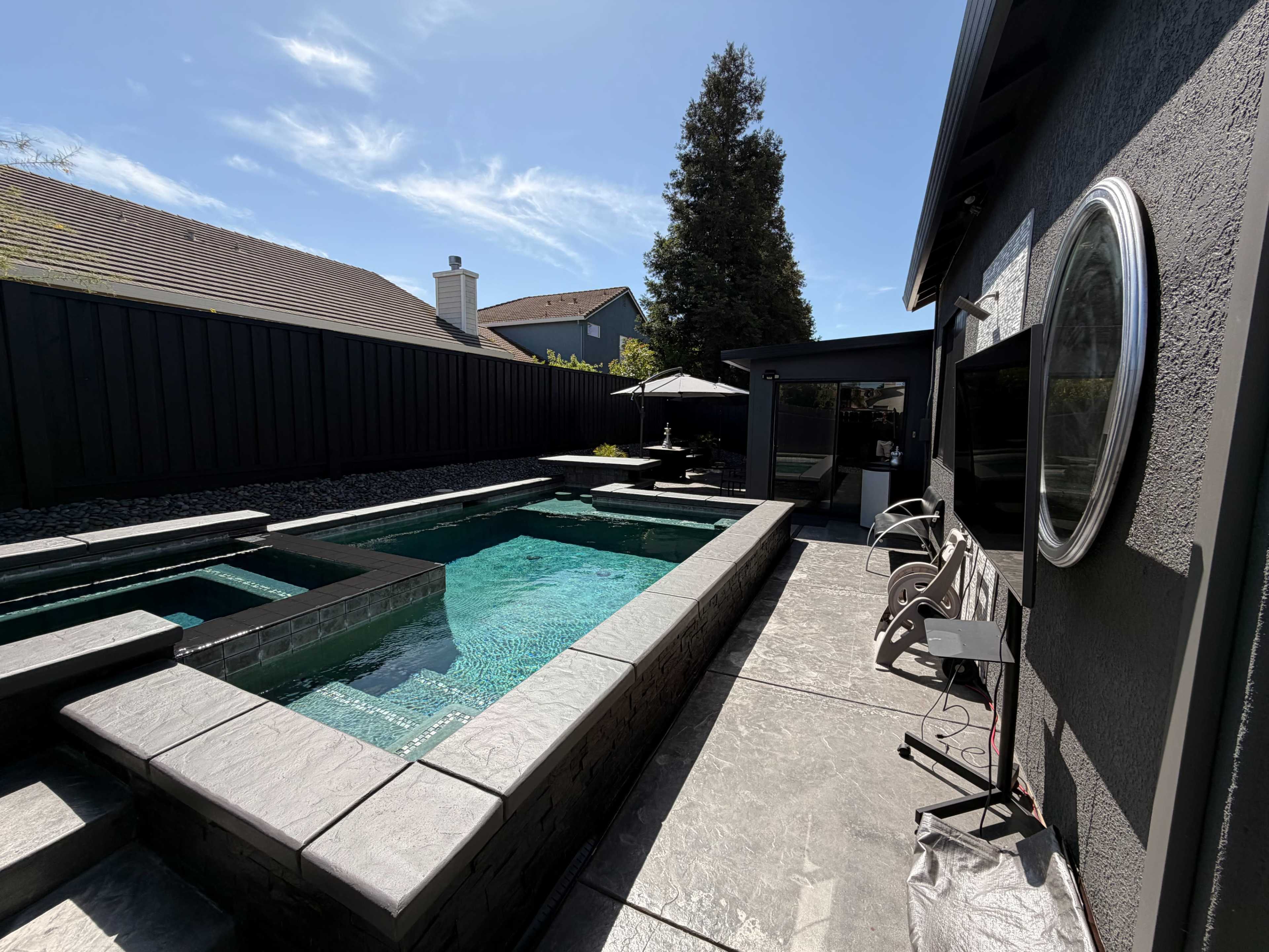 The image shows a modern backyard with a swimming pool, spa, and seating area adjacent to a house.
