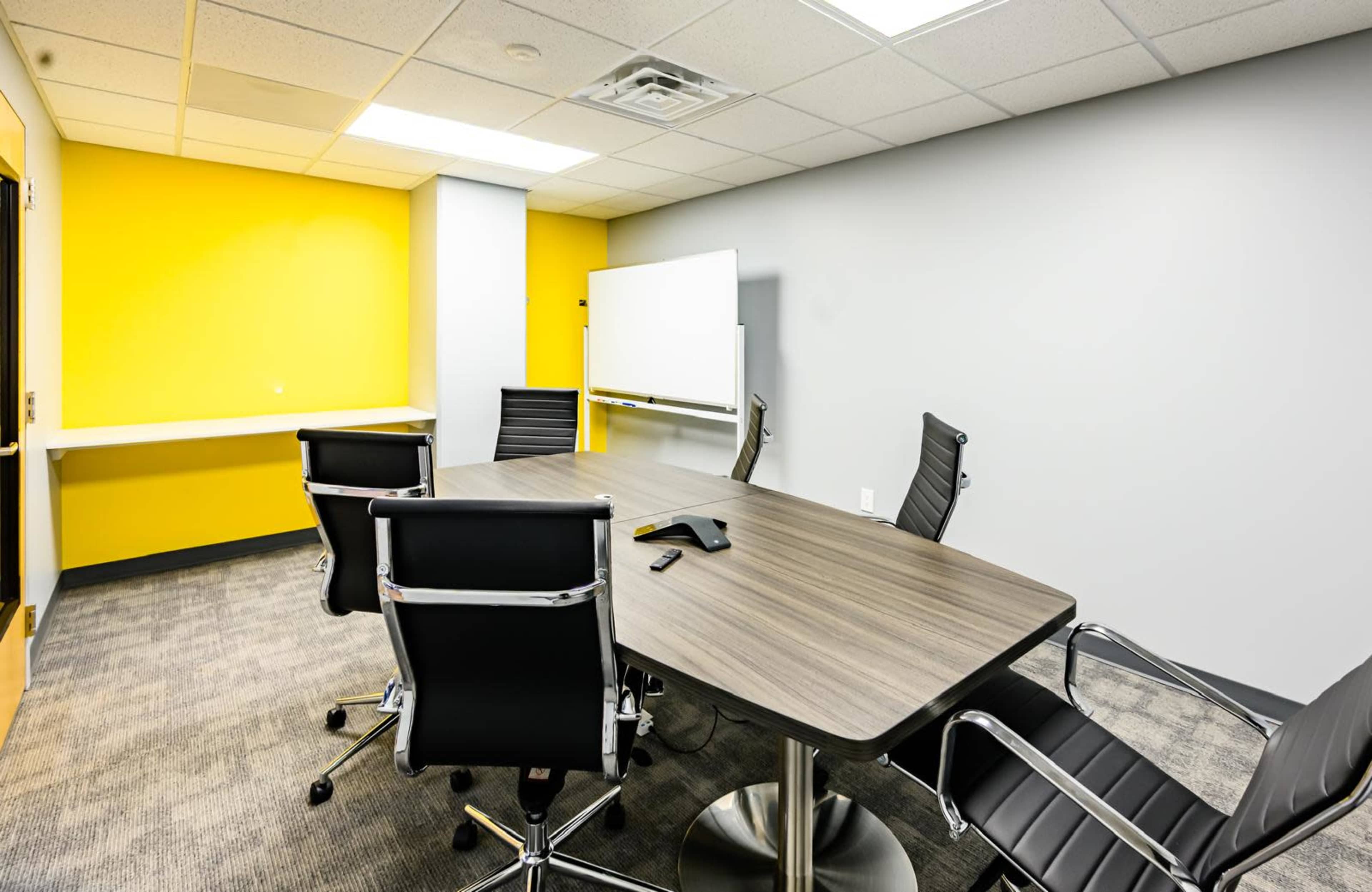 A conference room features a wooden table surrounded by four black chairs, with a whiteboard and a yellow wall in the background.