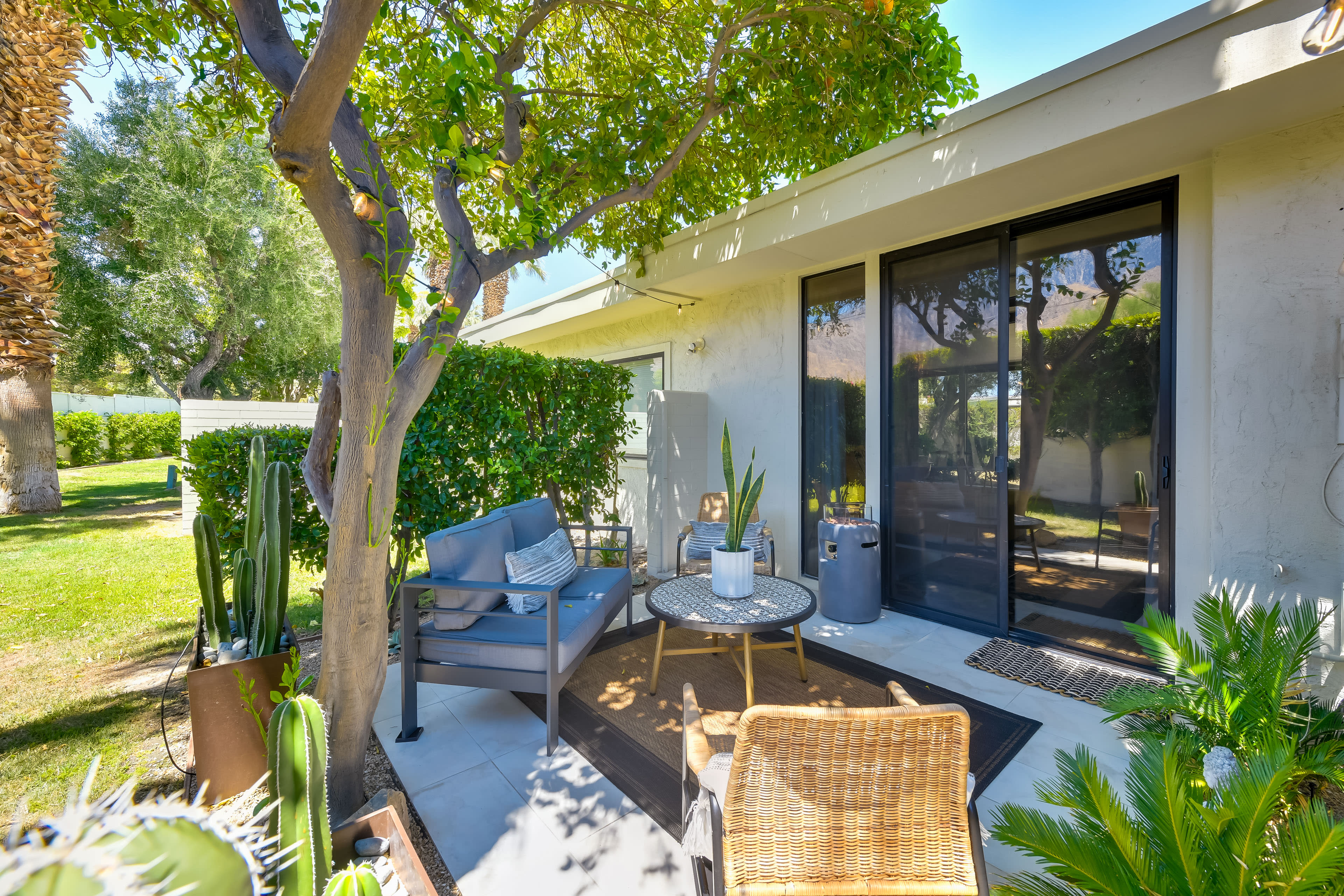 The image shows a landscaped patio area with comfortable seating, a table, and various plants surrounding a modern building.
