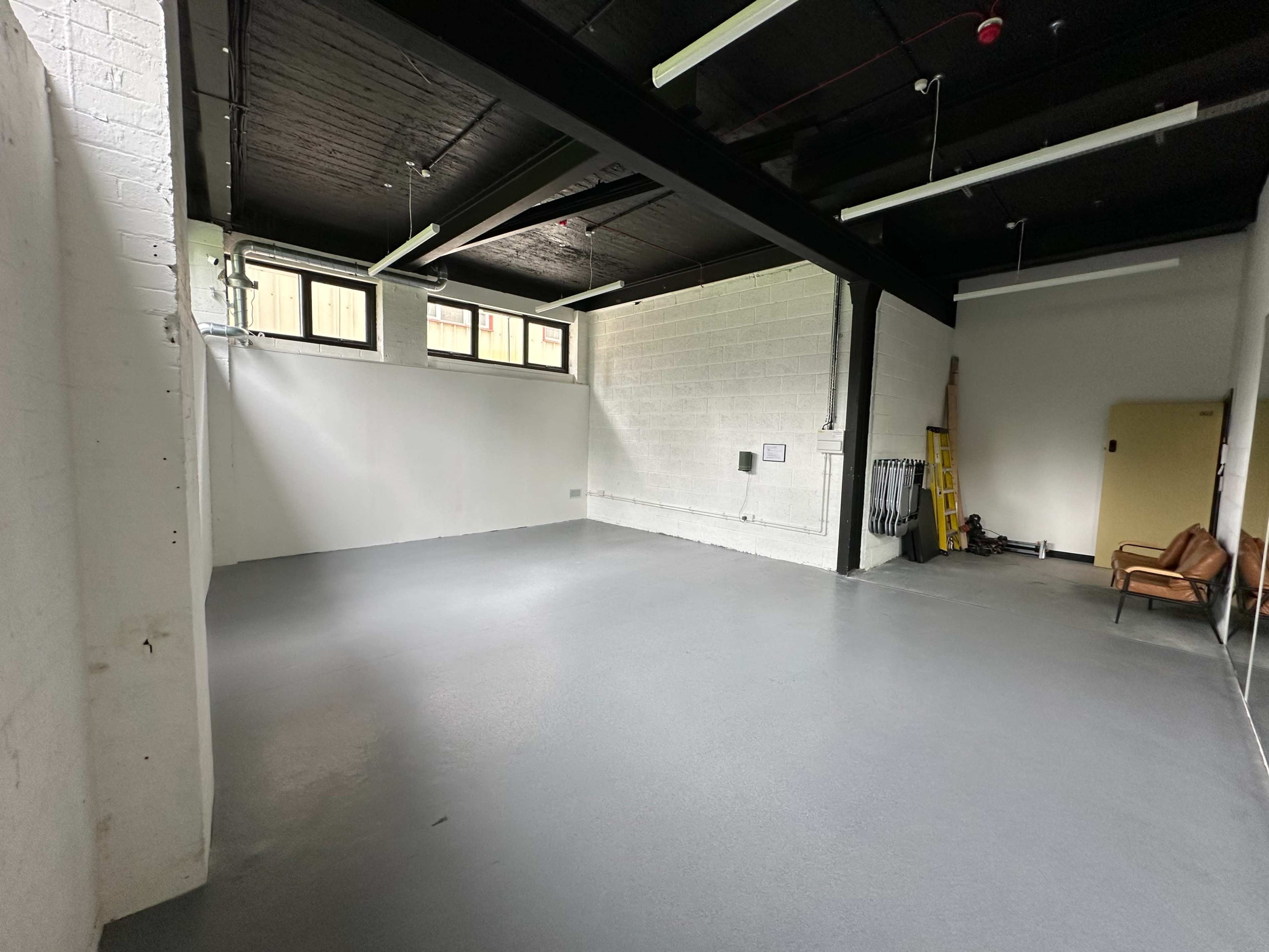 A spacious, empty room with a concrete floor, white walls, and large windows, featuring a chair and some equipment against one wall.