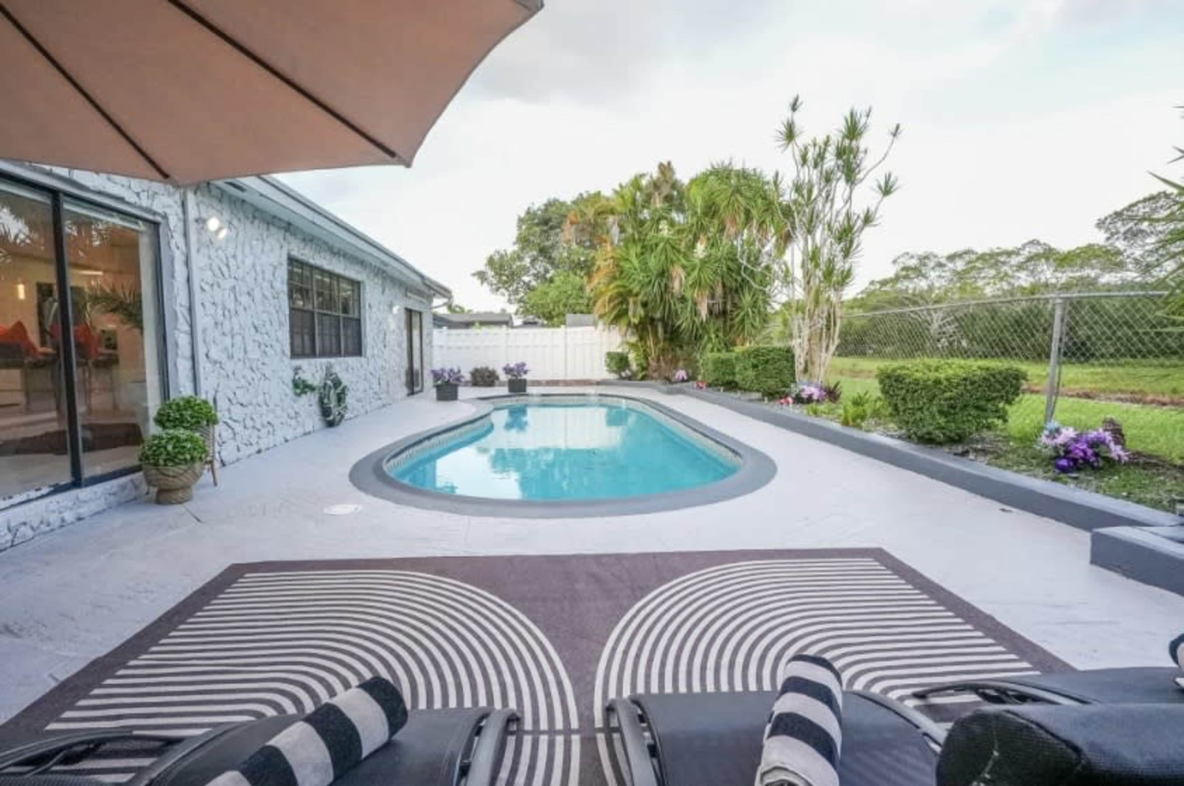 The image shows a backyard pool area with lounge chairs, surrounded by landscaped plants and a privacy fence.