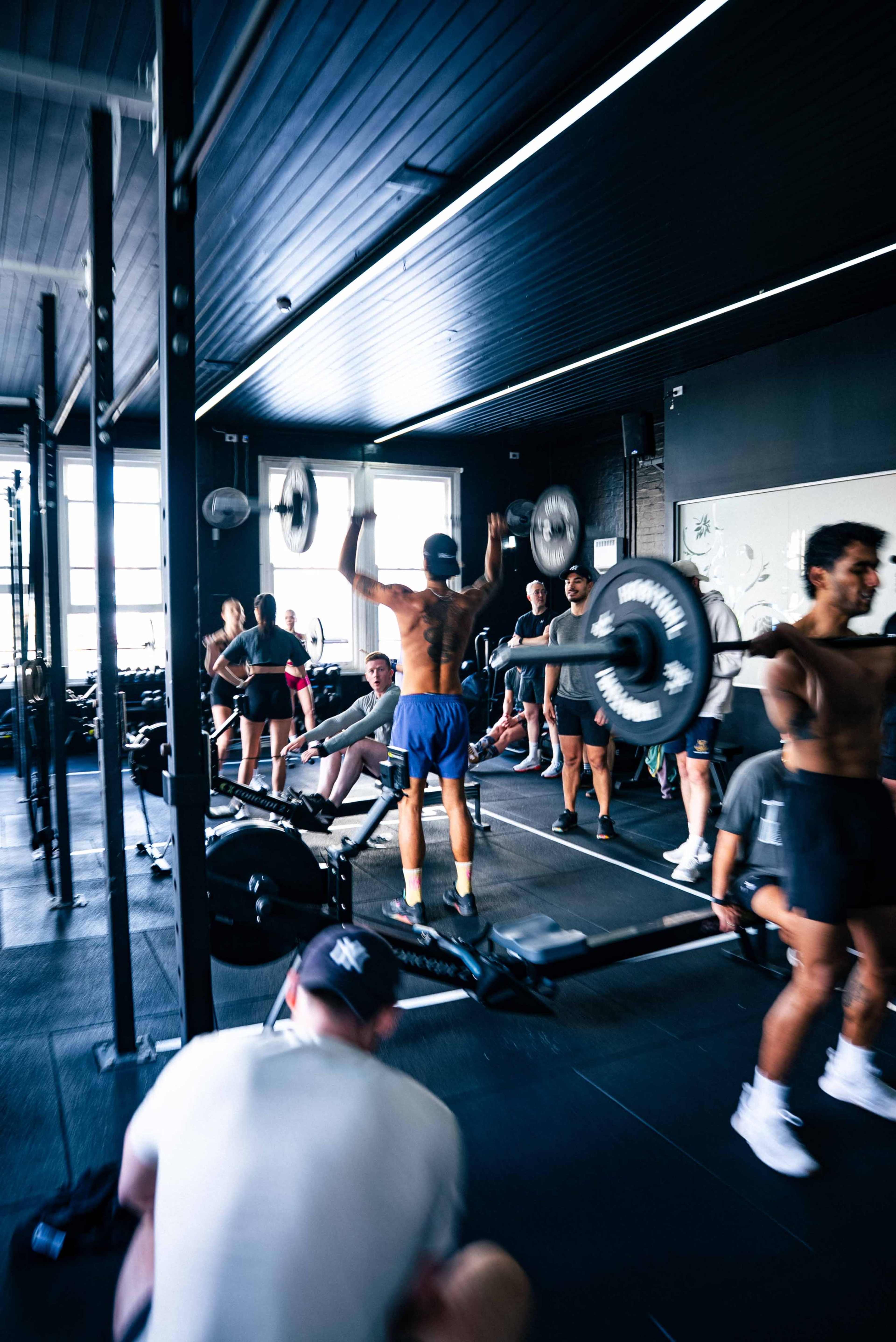 A group of individuals exercises in a gym, with some lifting weights and others using rowing machines.