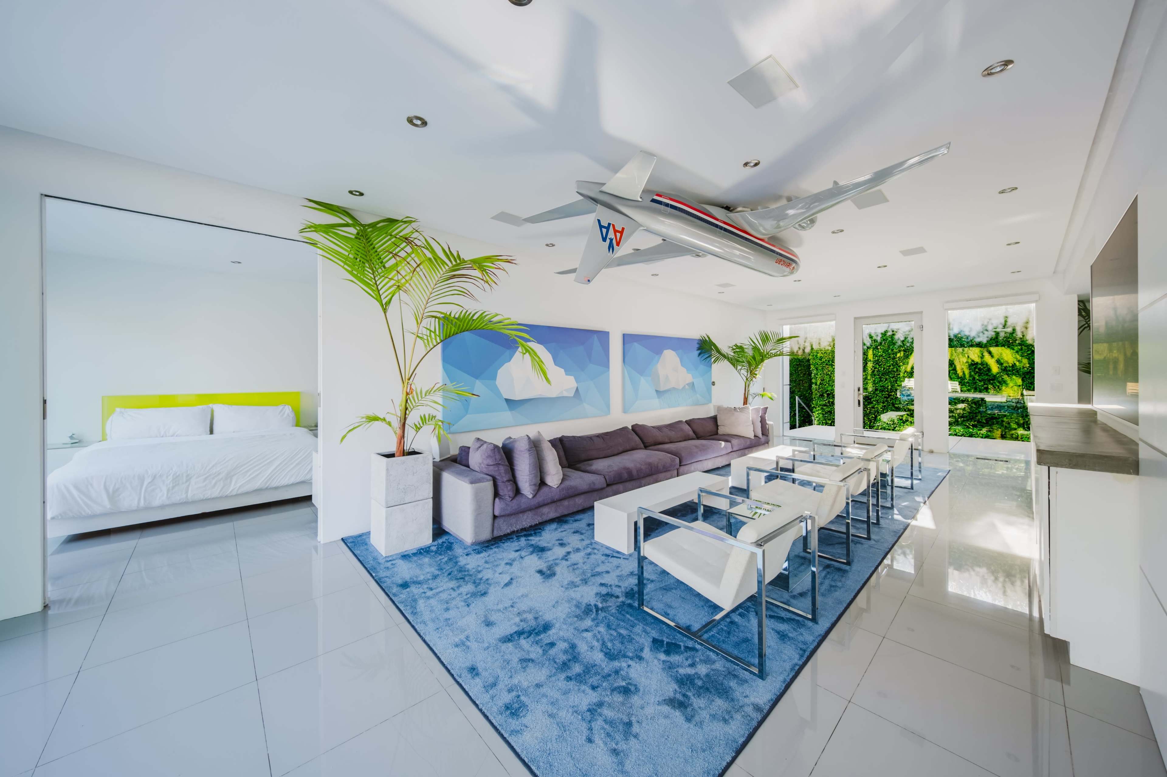 The image shows a modern, open-concept living area with a large purple sofa, several glass-top tables, and a model airplane hanging from the ceiling, adjacent to a bedroom with a white bed visible through an open doorway.