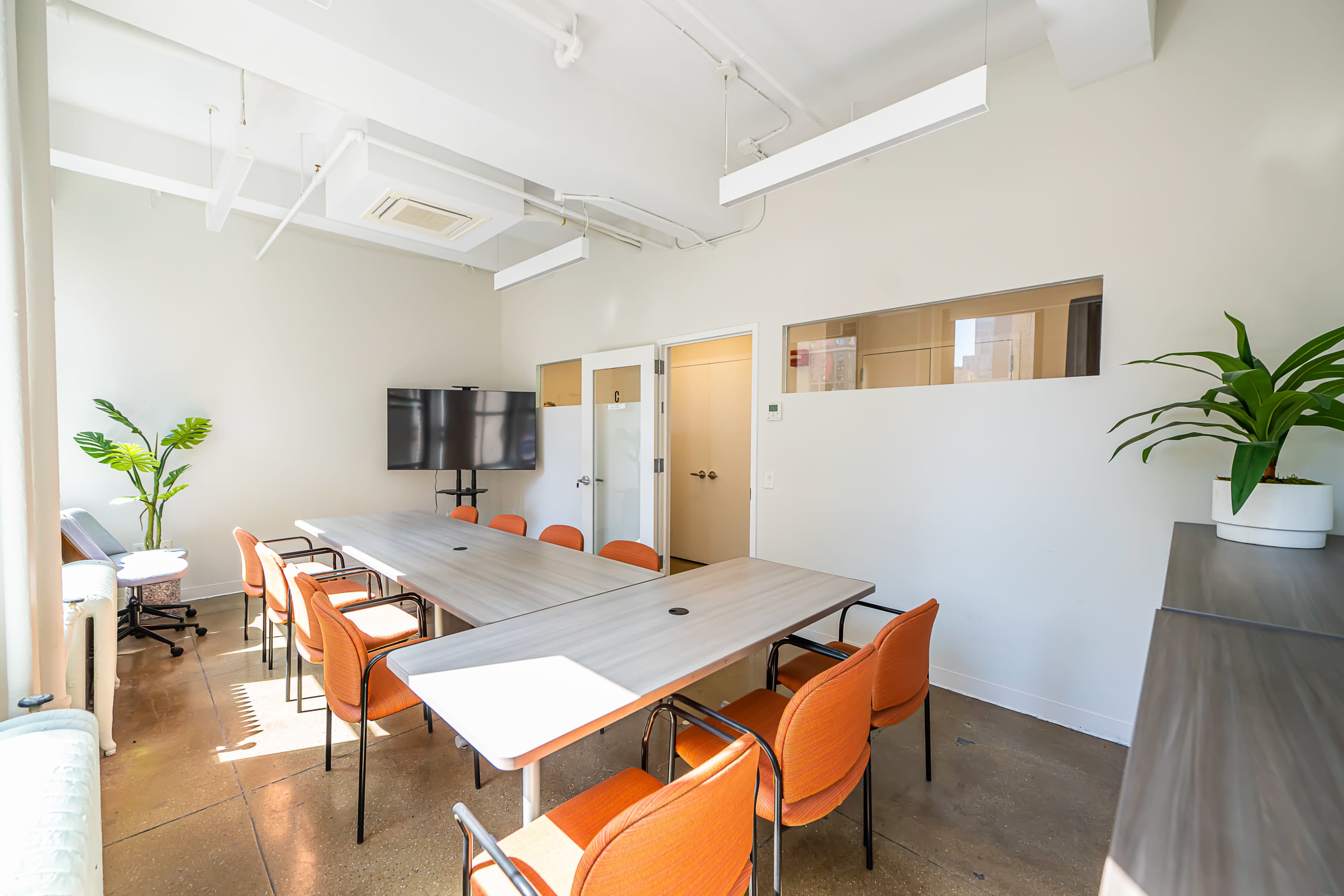 A modern conference room features a long wooden table surrounded by orange chairs, with a television screen mounted on the wall and a plant in the corner.