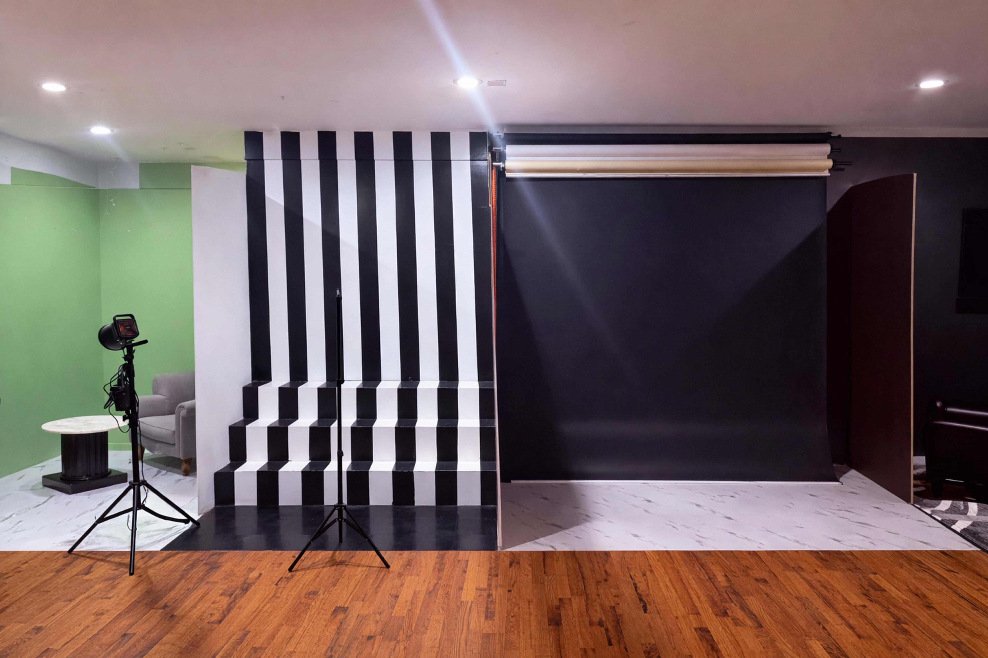 The image shows a photography studio featuring a black and white striped backdrop, a solid black backdrop, and a green wall, along with lighting equipment set up in front of the scenes.