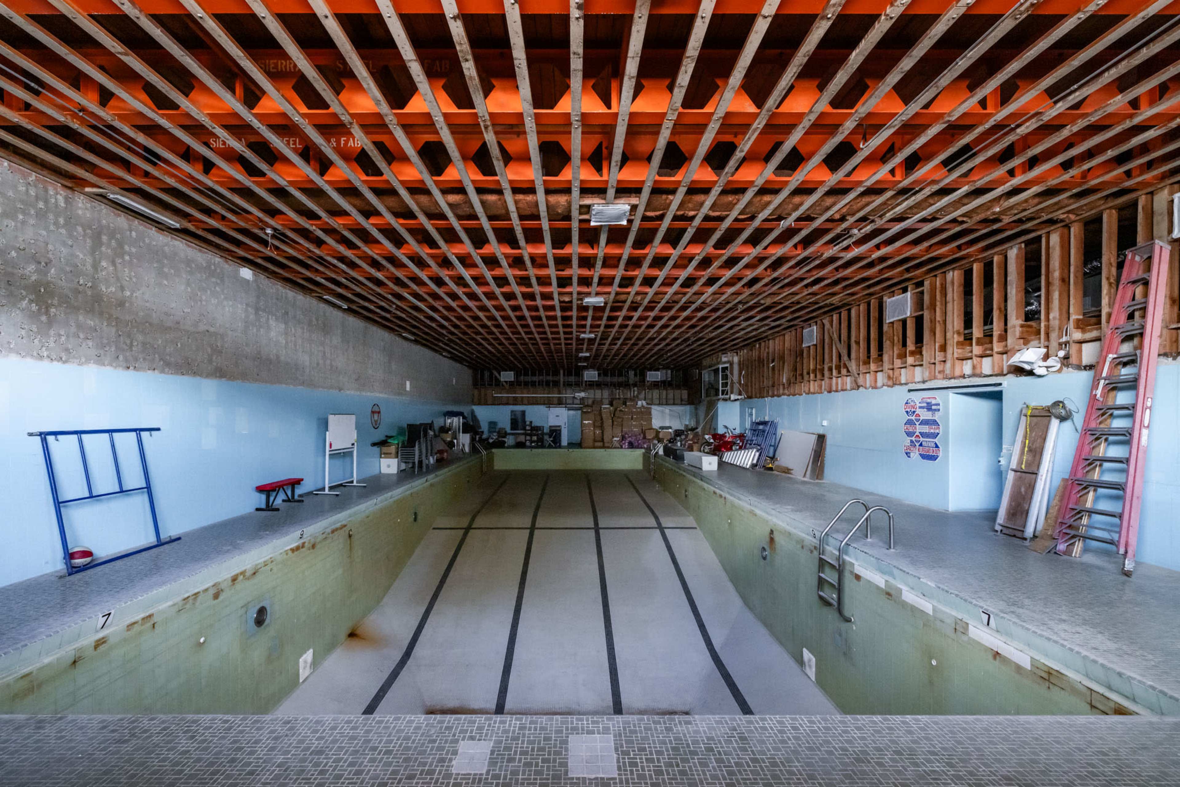 An empty indoor swimming pool with a partially exposed wooden ceiling and storage items along the sides.