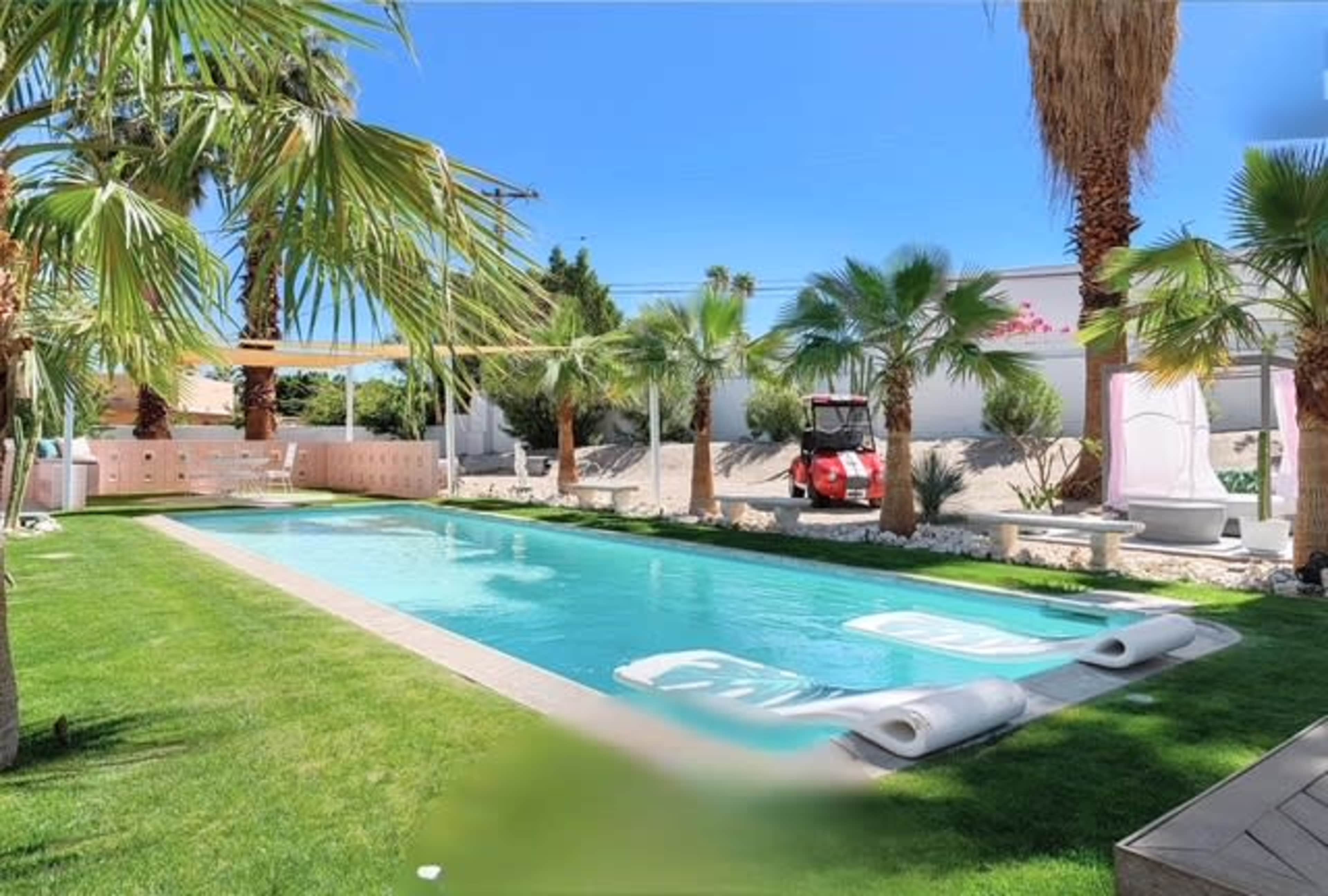 The image shows a swimming pool surrounded by palm trees and lush green grass, with lounge chairs positioned on one side.
