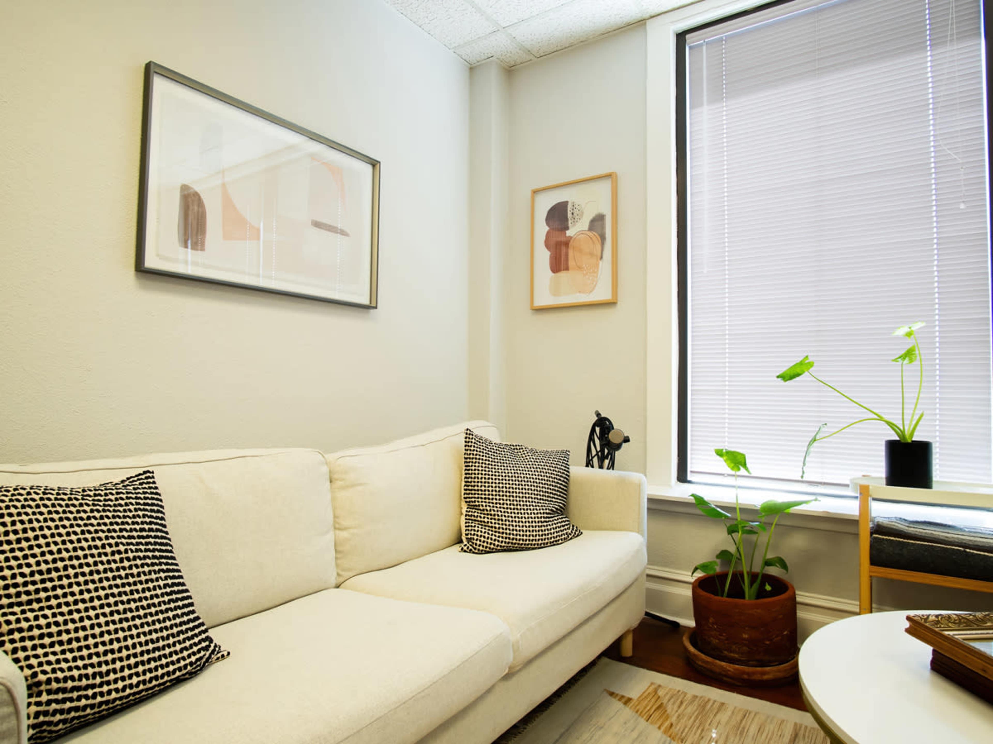 A light-colored sofa with two patterned pillows is positioned against a wall, accompanied by framed artwork and a potted plant in a window nook.
