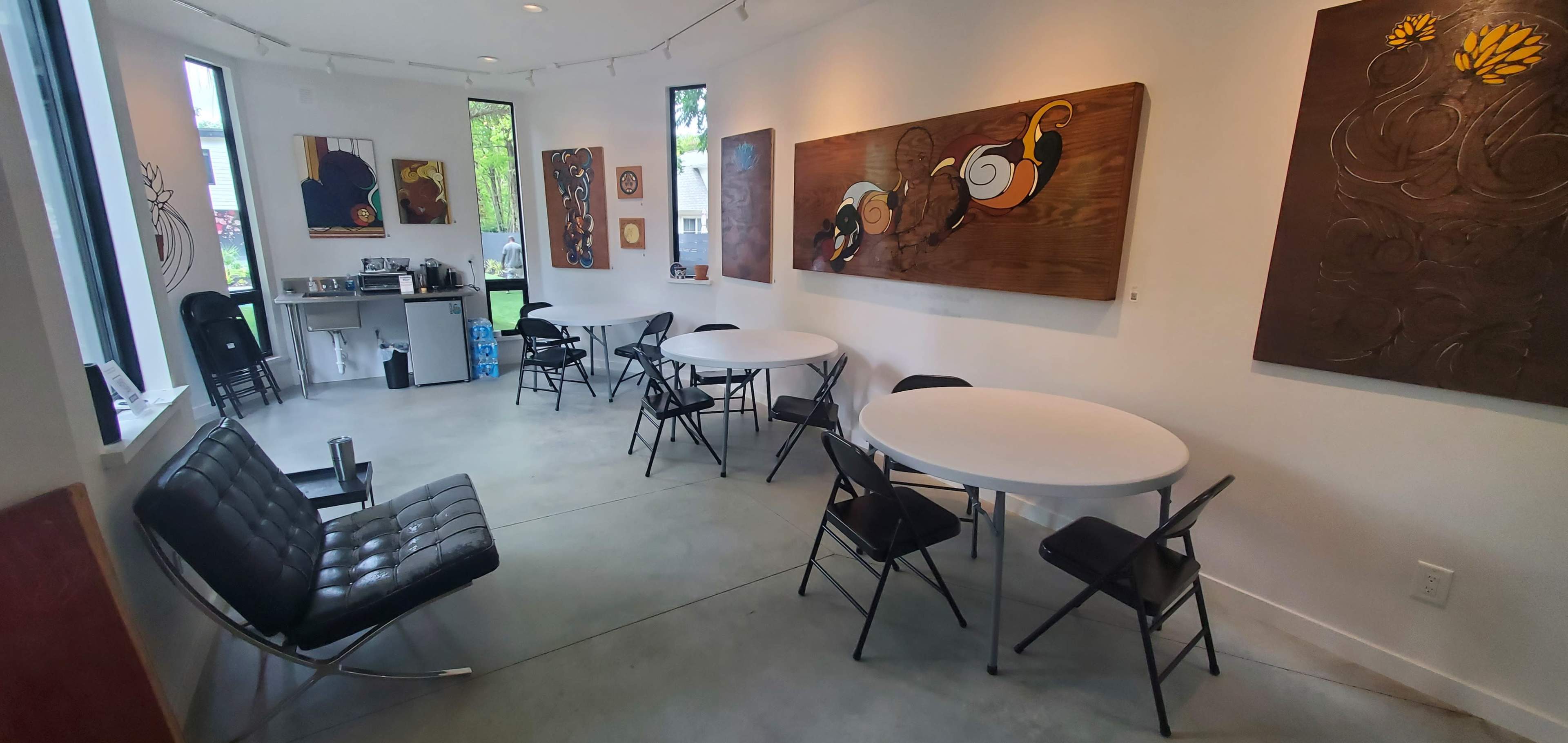 An art gallery with several wooden artworks on the walls, round tables and folding chairs arranged in the space, and a kitchenette area in the background.