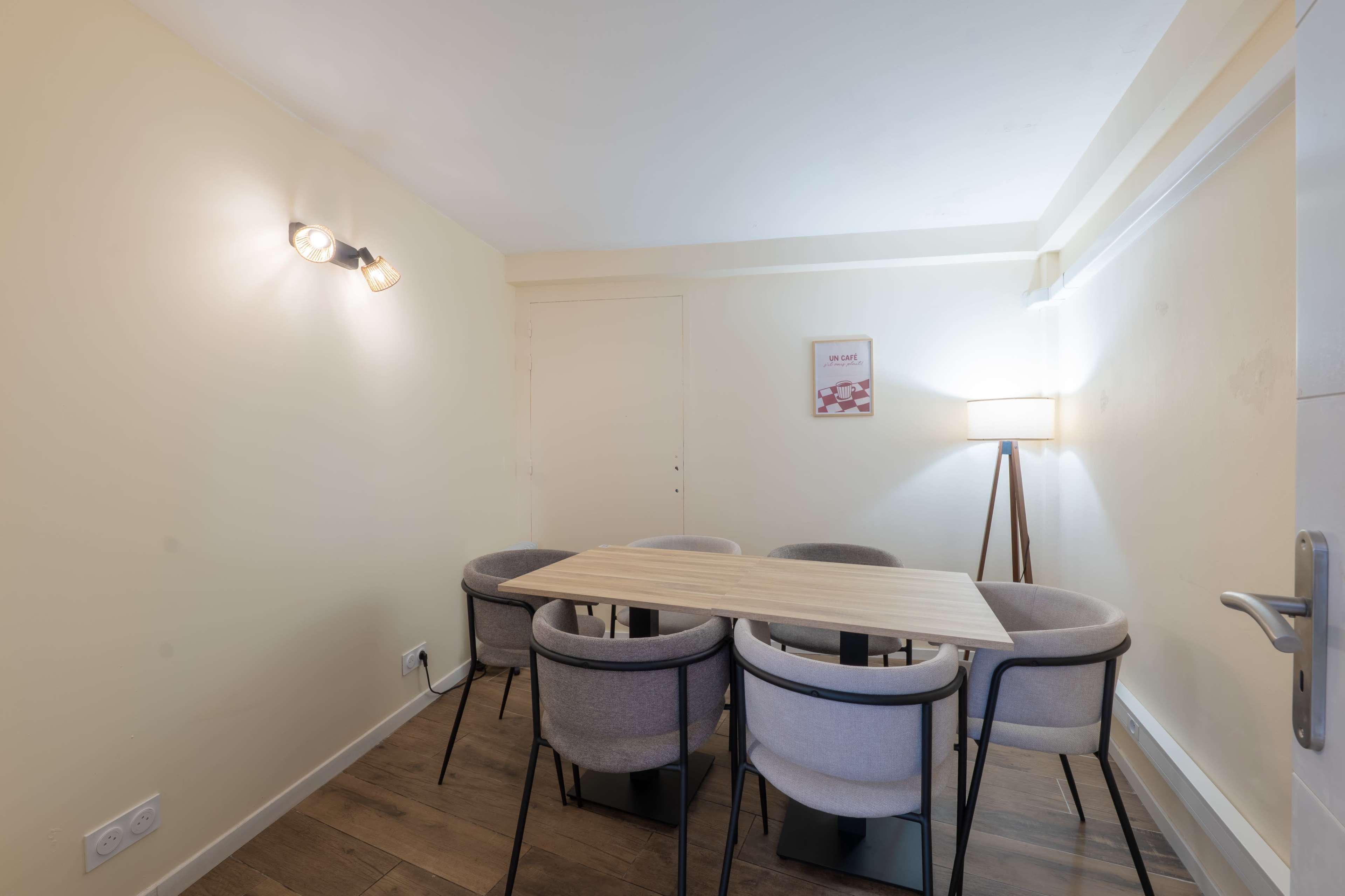 The image shows a small dining room with a wooden table surrounded by six gray chairs, a floor lamp, and a wall light, against a pale yellow backdrop.