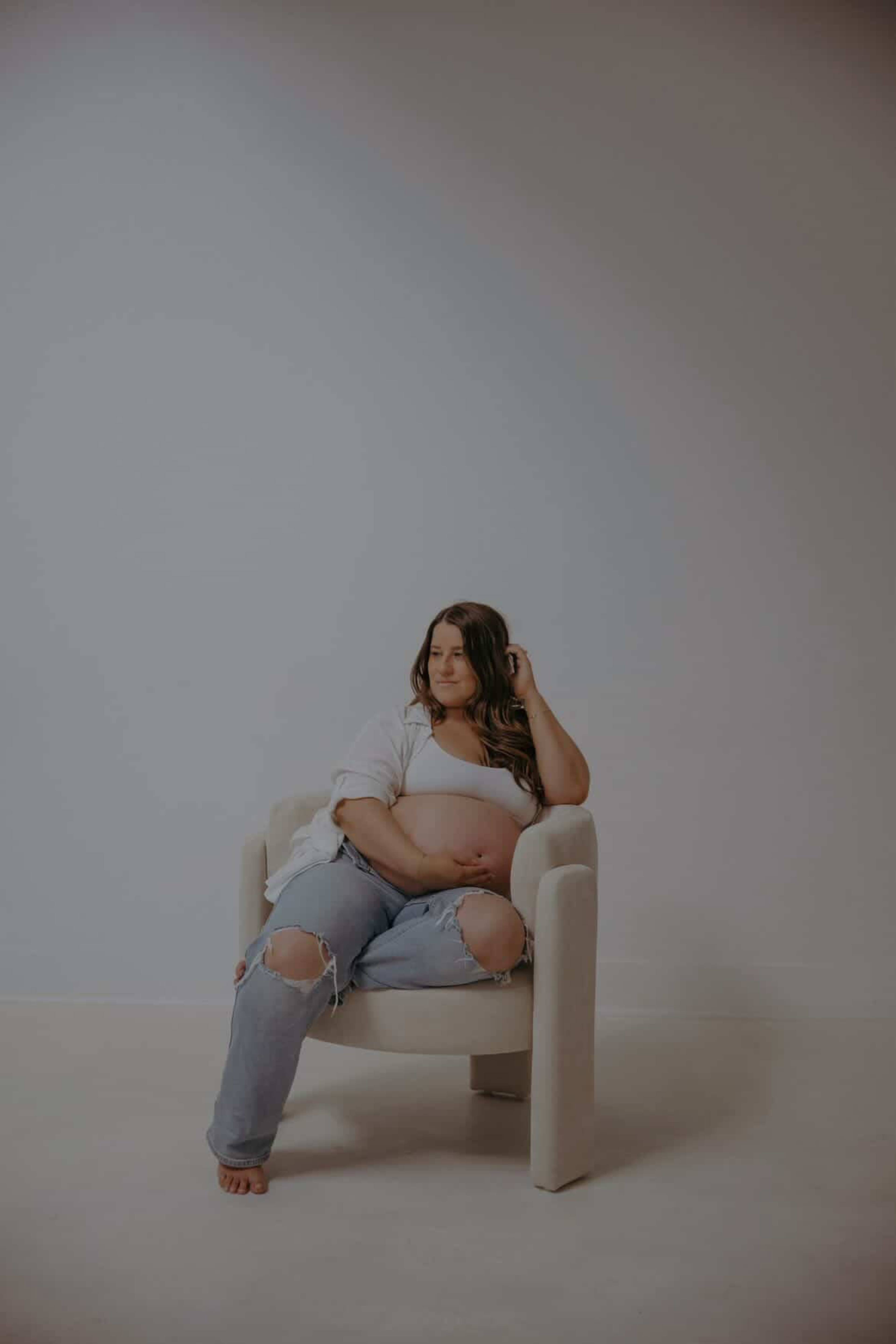 A person is sitting on a white armchair against a light-colored background, wearing a casual outfit with ripped jeans.