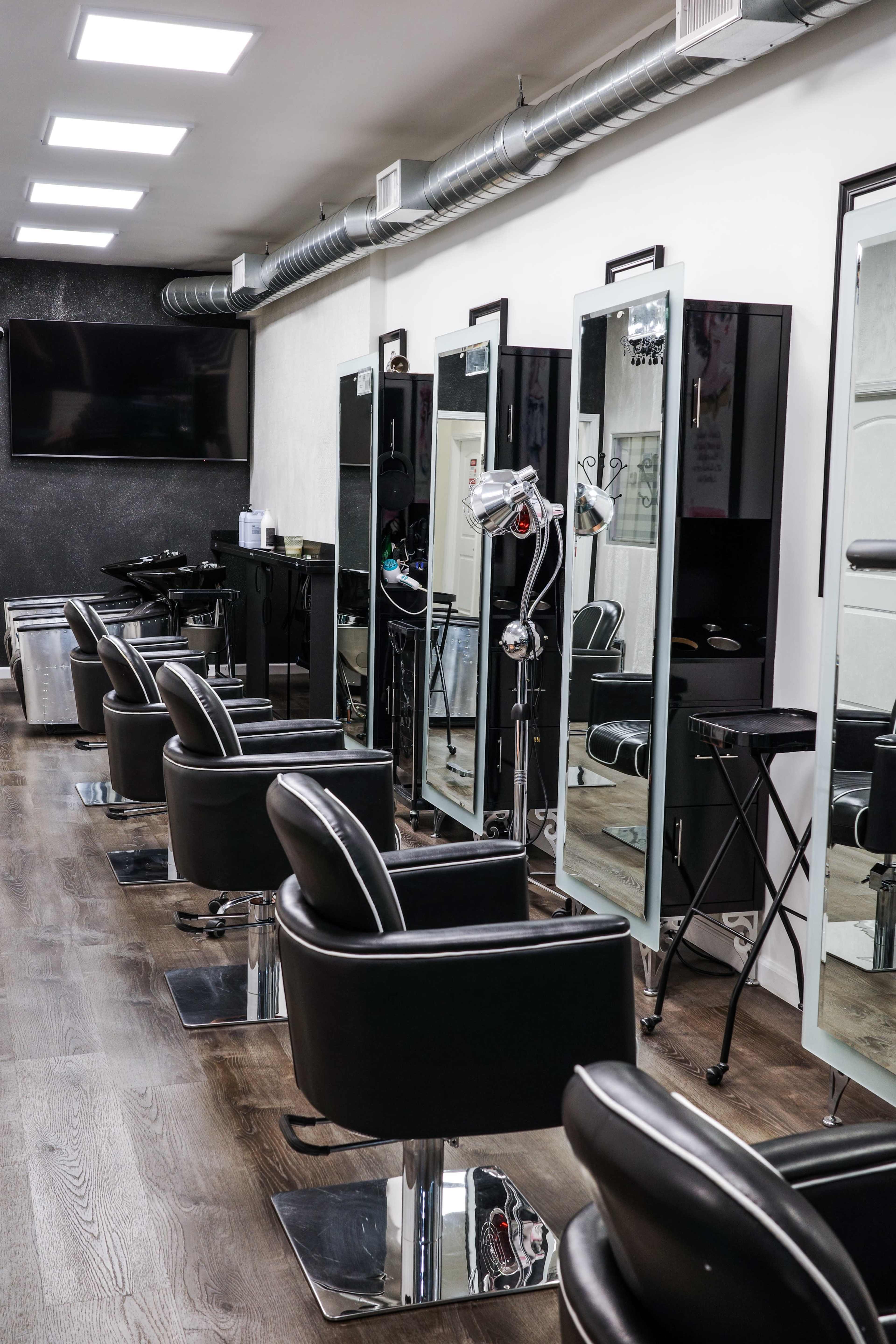 A modern hair salon features several styling stations with large mirrors and black chairs arranged along a well-lit wall.