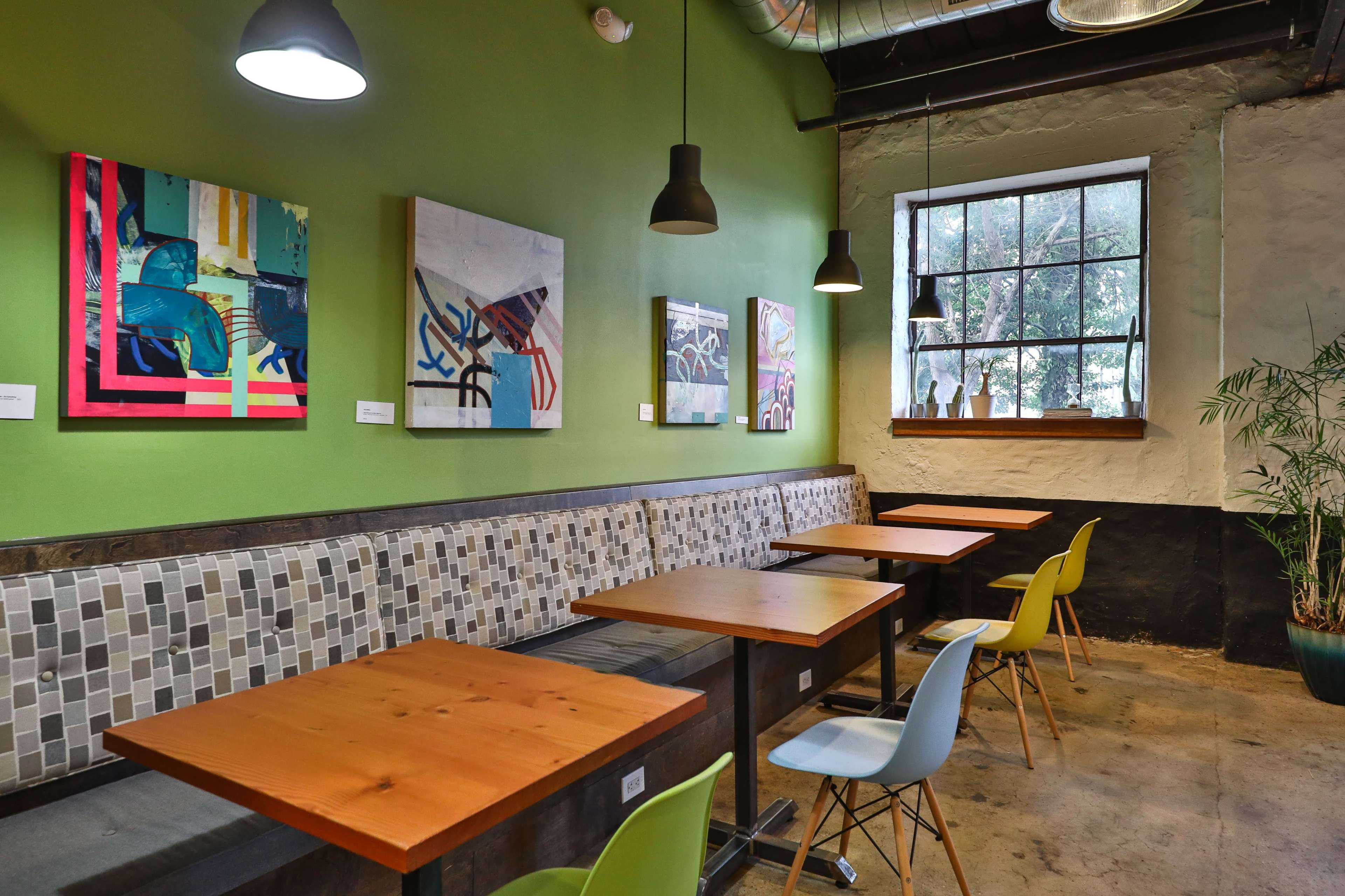 A cozy café interior featuring green walls, several artworks on display, wooden tables, and a large window with potted plants.