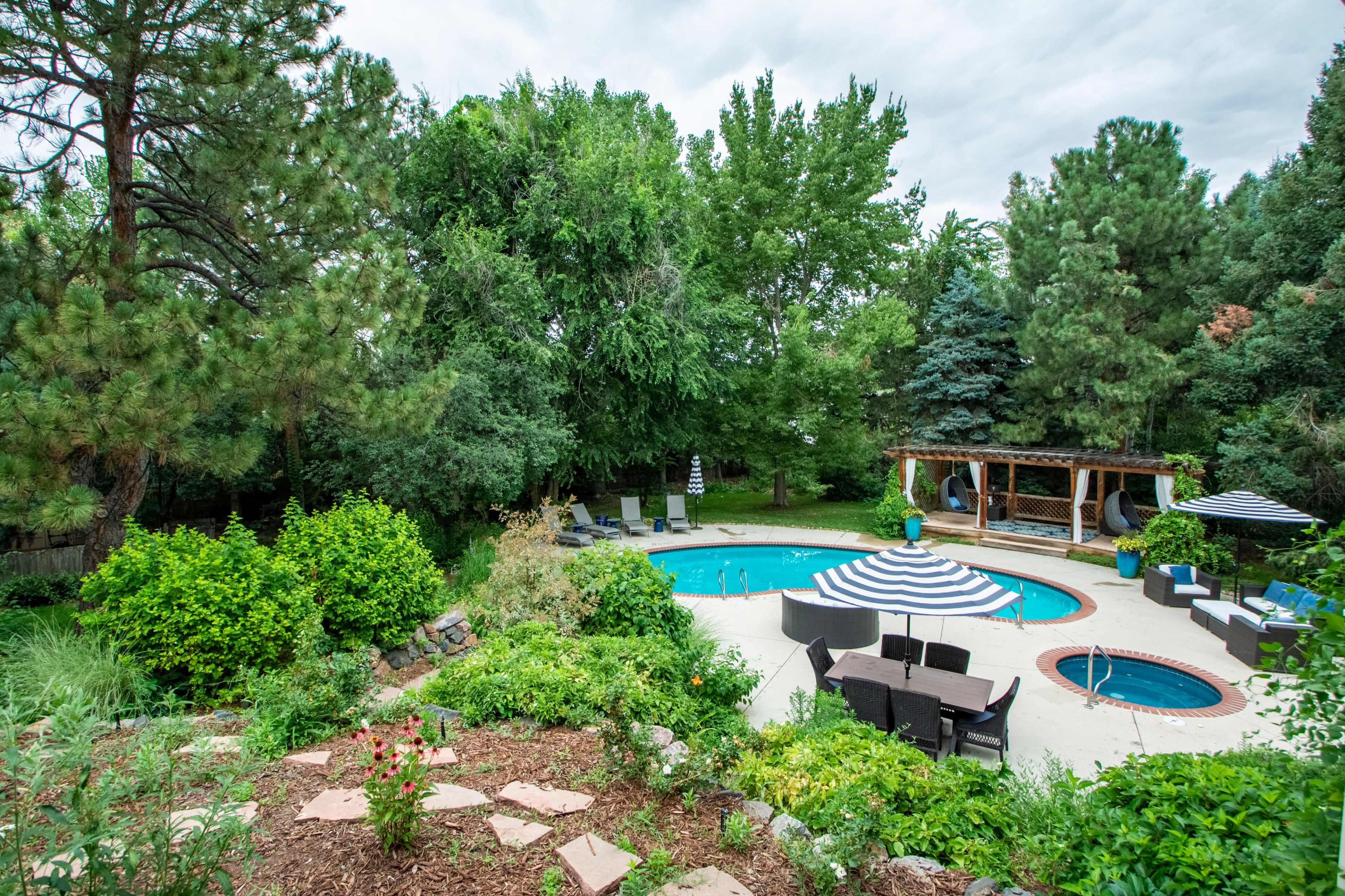 The image shows a landscaped backyard with a swimming pool, surrounded by trees and green shrubs, featuring a seating area and a gazebo with striped umbrellas.