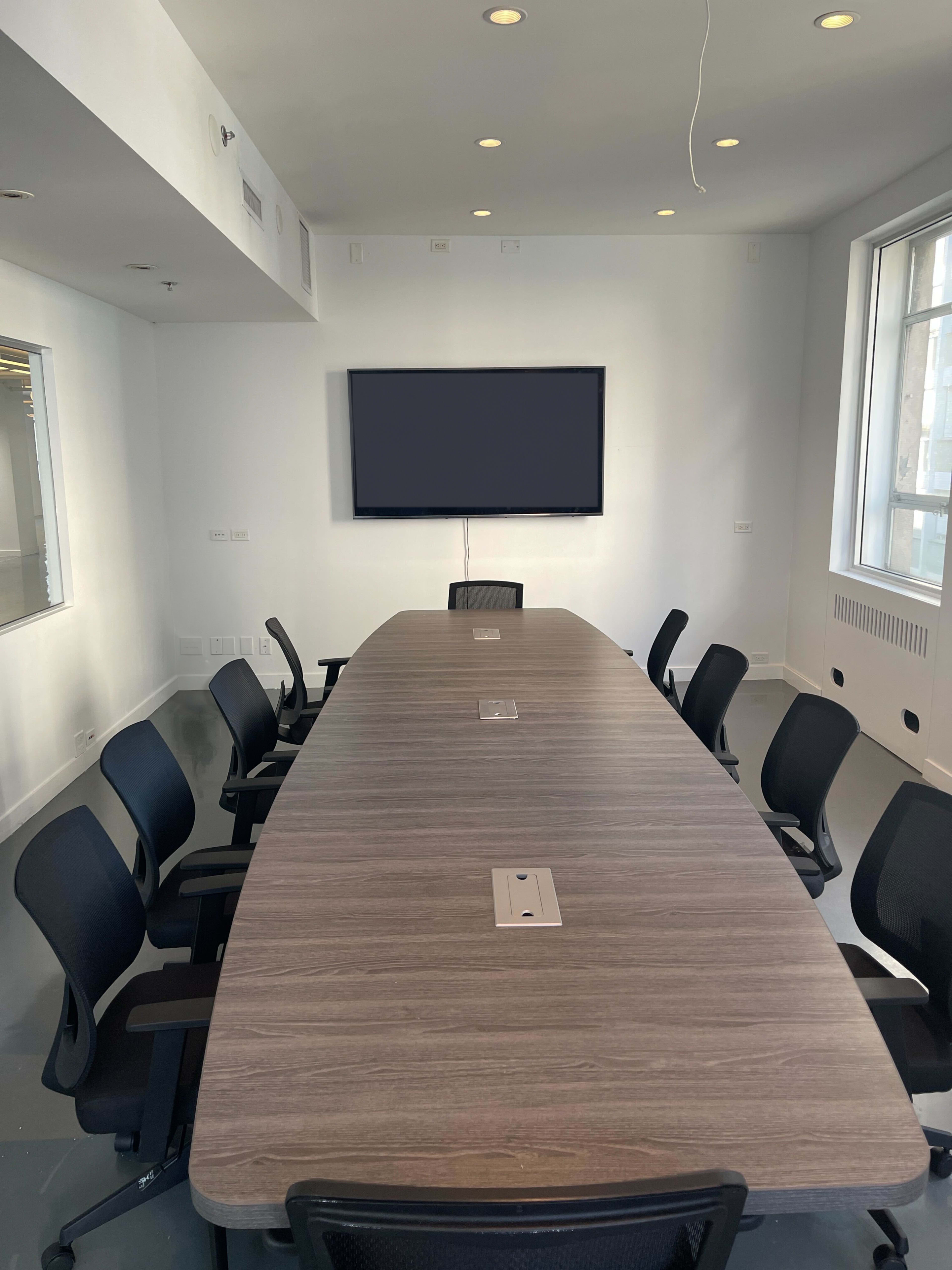 A large, oval conference table is surrounded by black ergonomic chairs in a brightly lit meeting room, featuring a wall-mounted screen and windows.