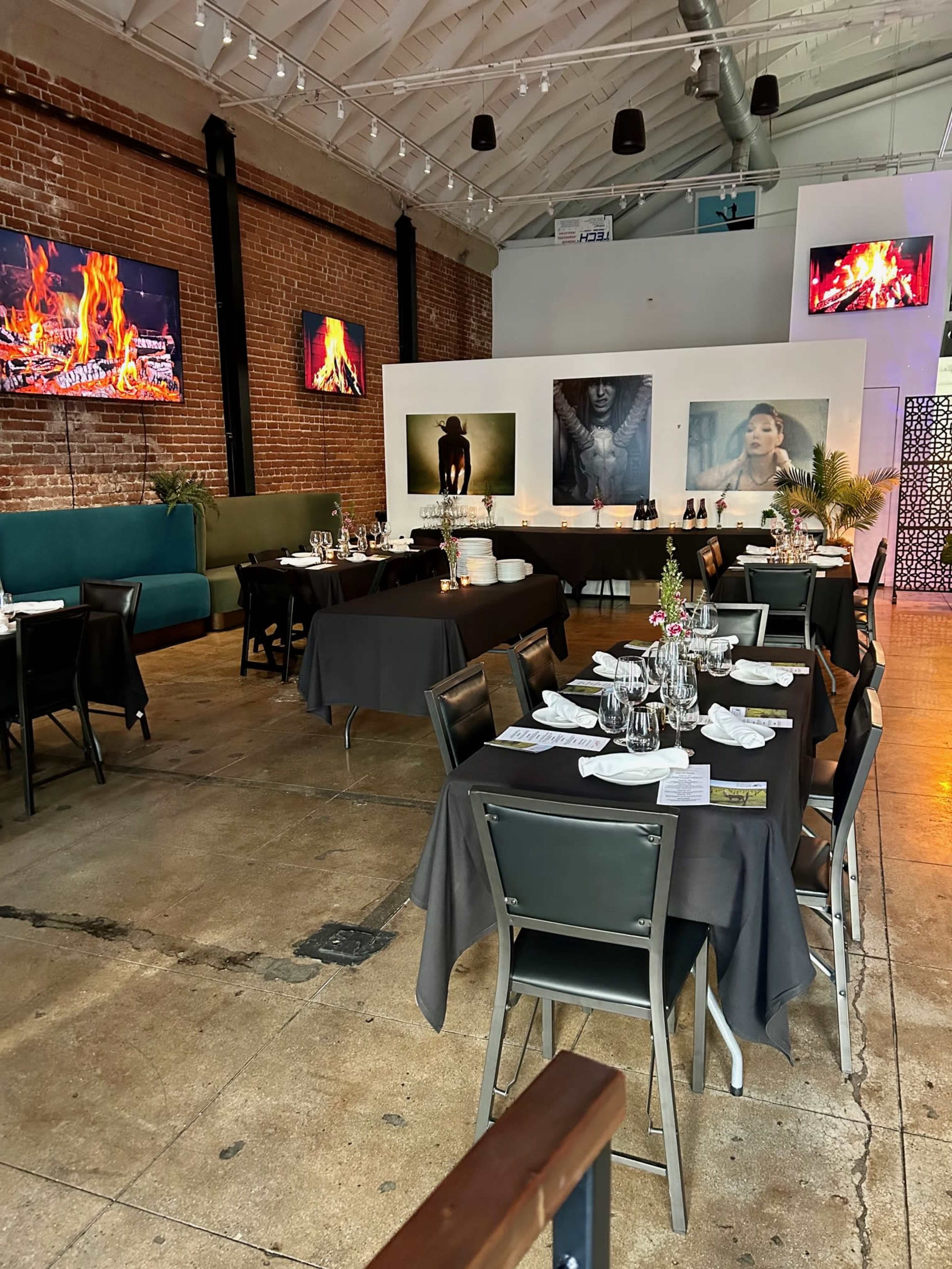 The image shows an indoor restaurant space with dark tables set for dining, a brick wall, and large screens displaying fireplace visuals.