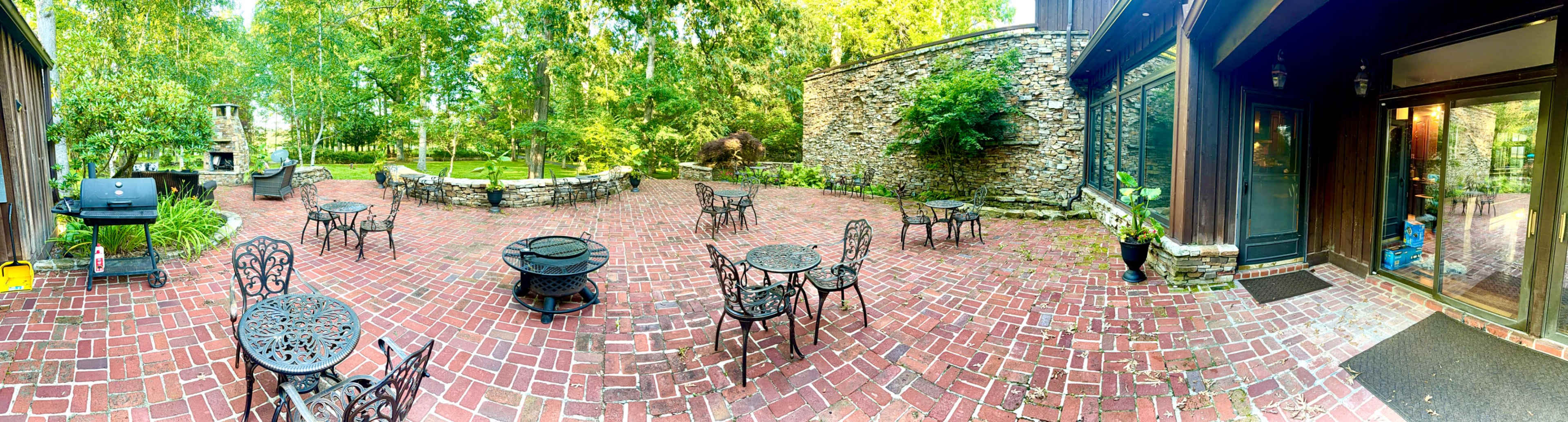 The image shows a brick patio with several metal tables and chairs surrounded by greenery and stone walls.