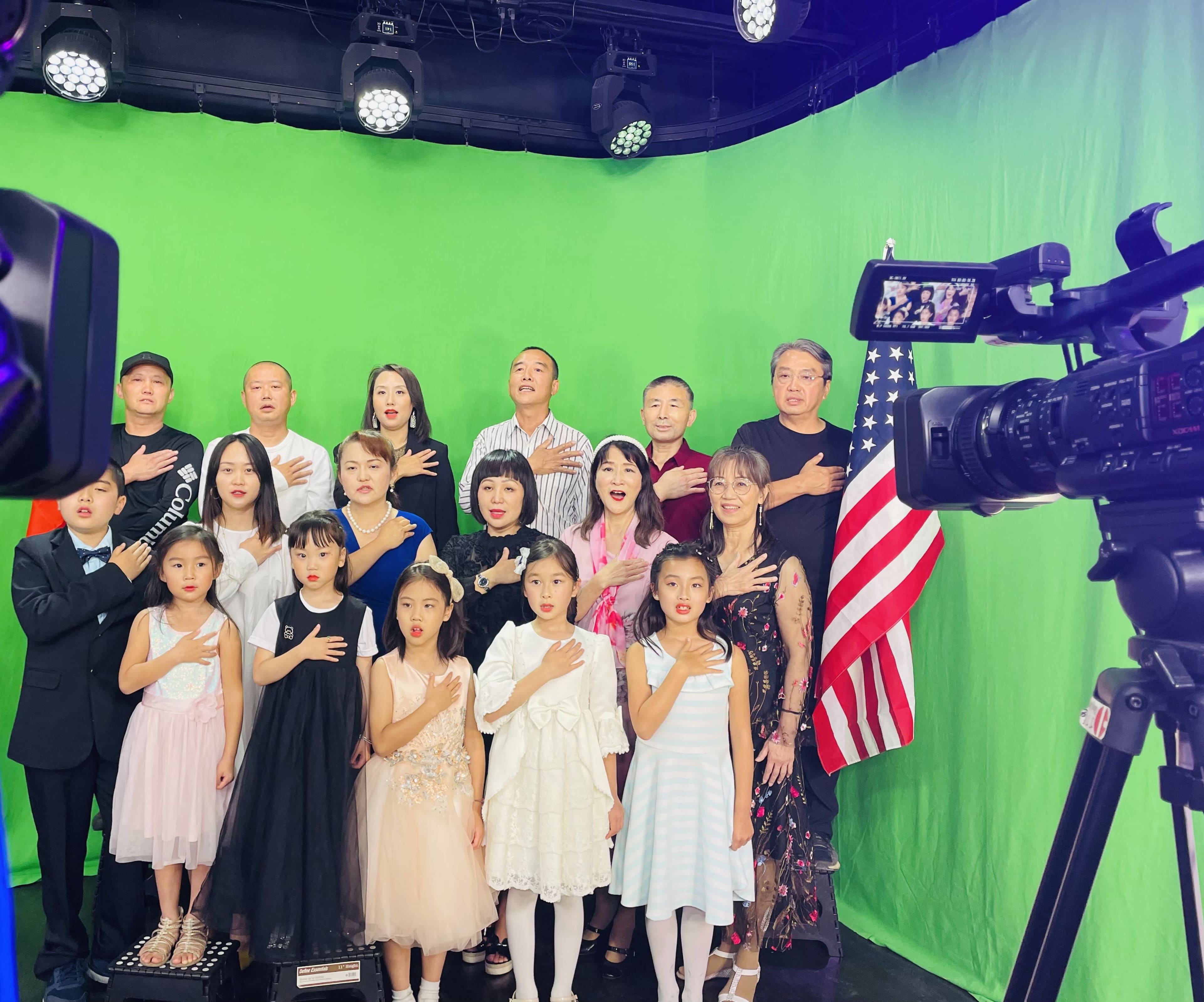 A group of adults and children stands in front of a green screen, with some members placing their hands over their hearts and an American flag on display.