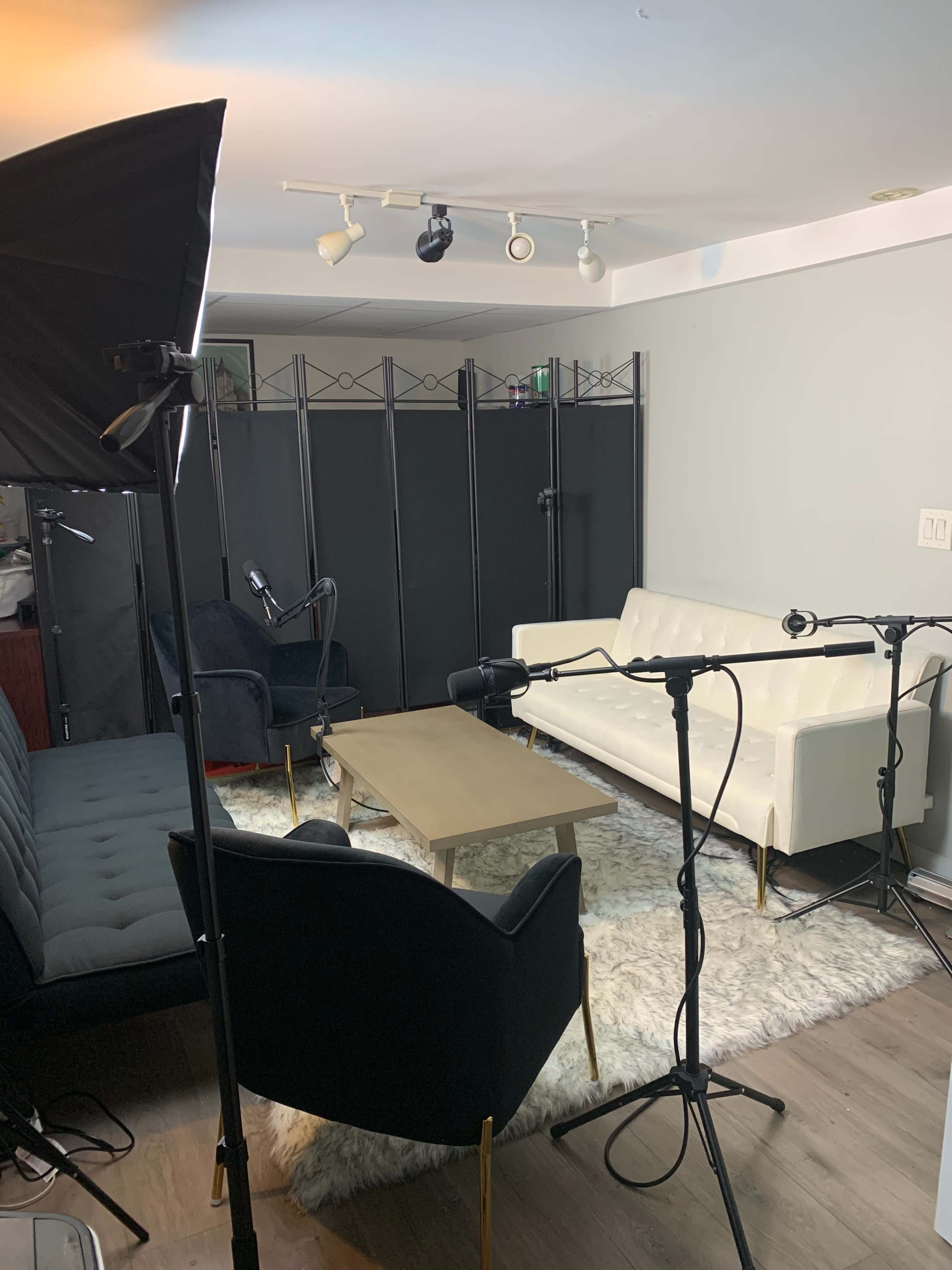 A small recording studio features a white sofa, a dark chair, a wooden coffee table, and microphones set up on stands, all placed on a plush rug in front of a folding screen.