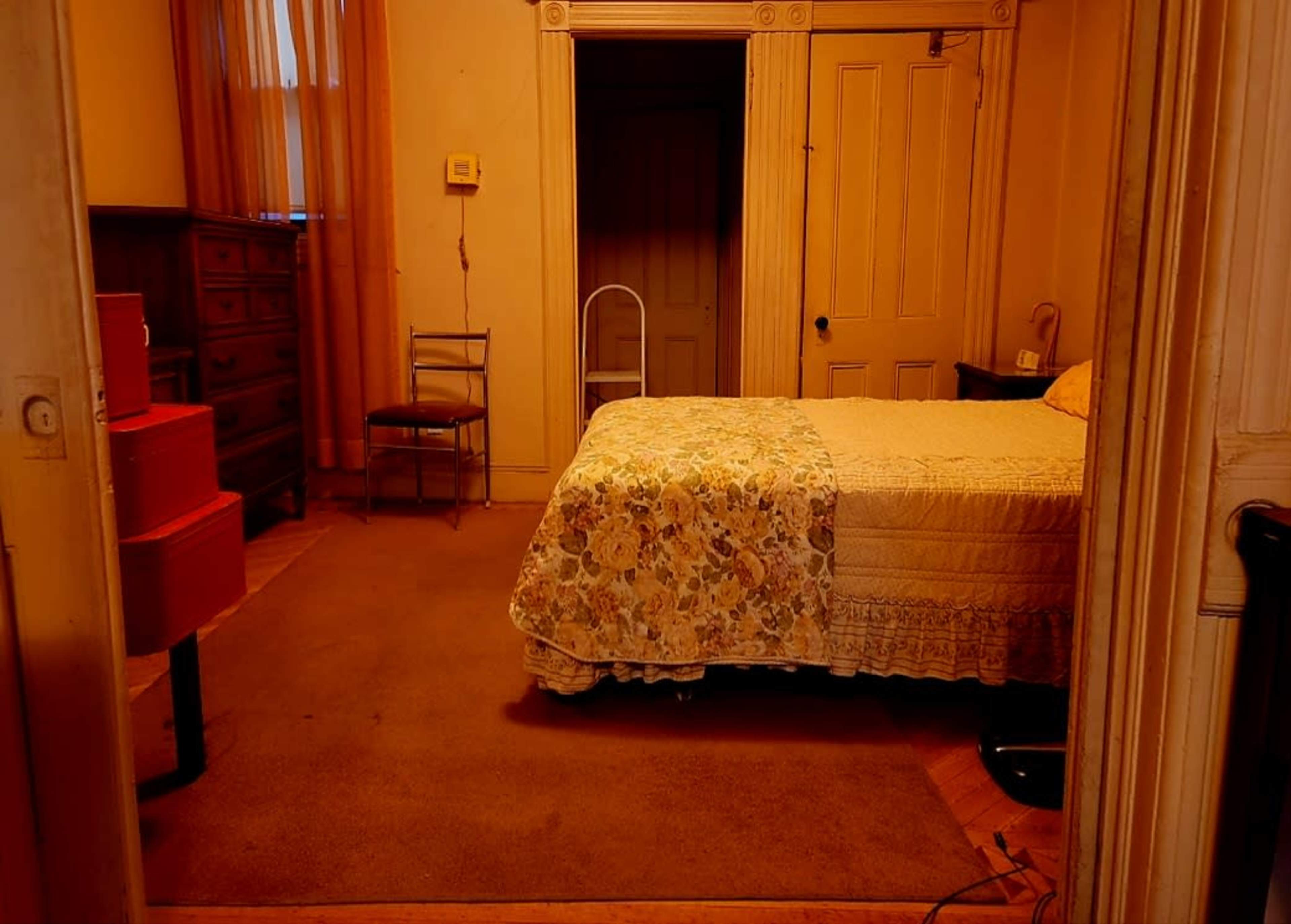 The image shows a simple bedroom with a bed covered in a floral quilt, a dresser, a chair, and a carpeted floor.