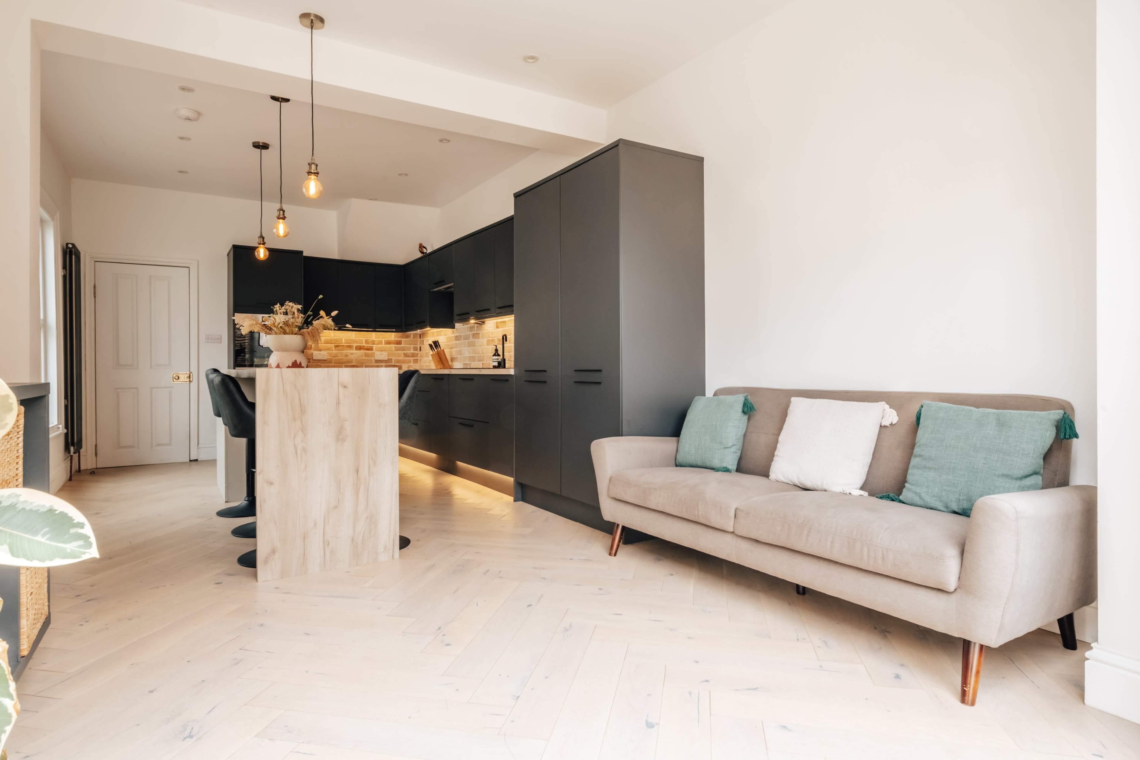 The image shows a modern kitchen and dining area with dark cabinetry, a wooden island, and a light-colored sofa in a well-lit space.