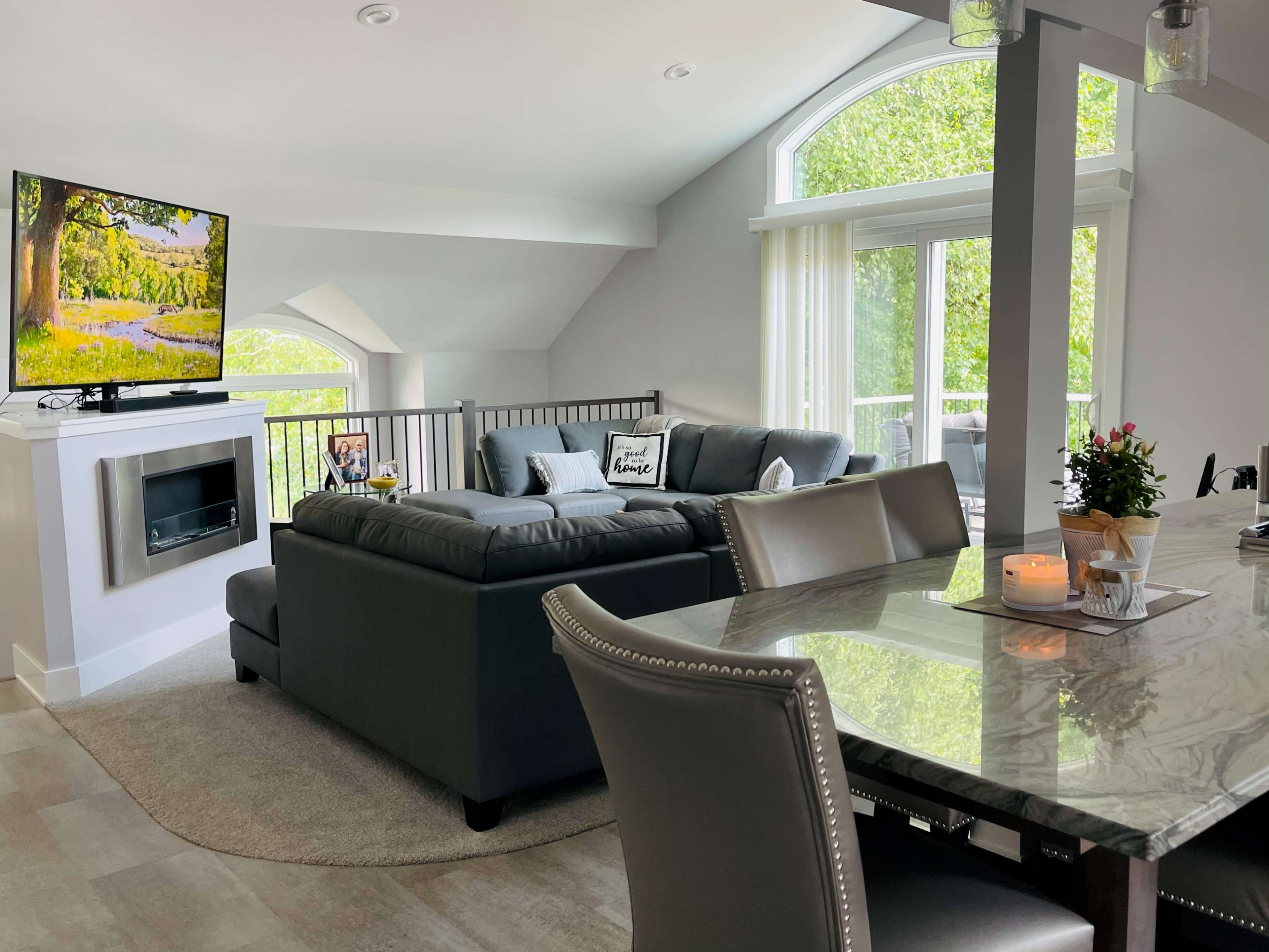 A modern living area features a gray sectional sofa facing a TV mounted above a decorative fireplace, with a dining table set nearby and large windows providing natural light.