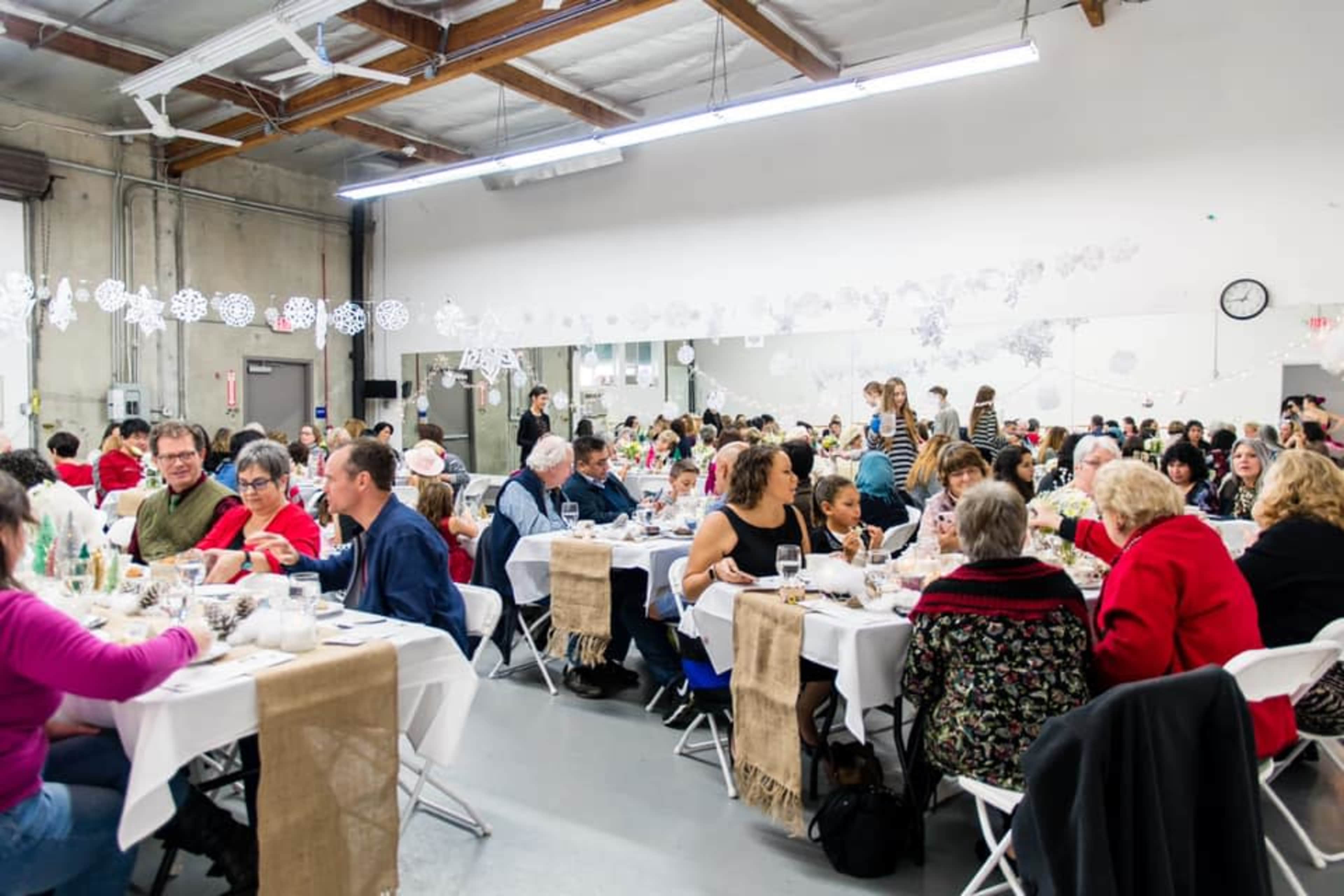 A large group of people sit at tables in a spacious hall, enjoying a meal together during a gathering.