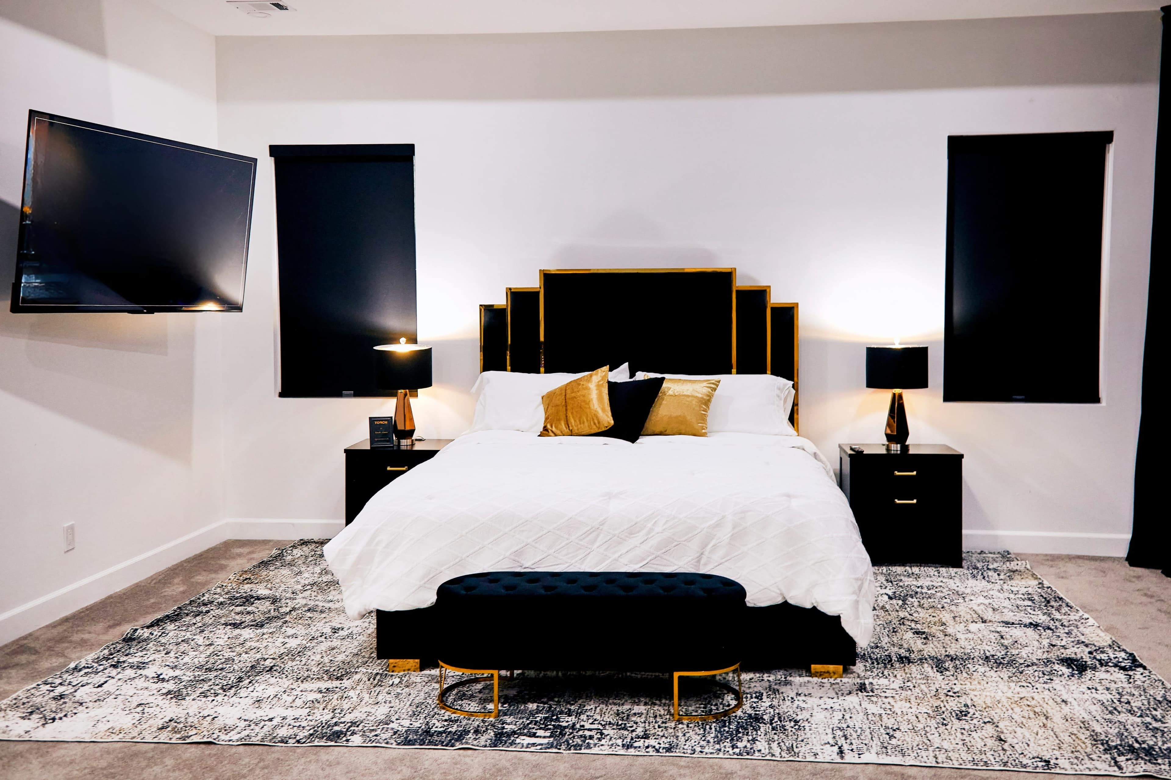 The image shows a modern bedroom featuring a queen-sized bed with a black and gold headboard, two bedside tables with lamps, a television mounted on the wall, and a patterned rug on the floor.