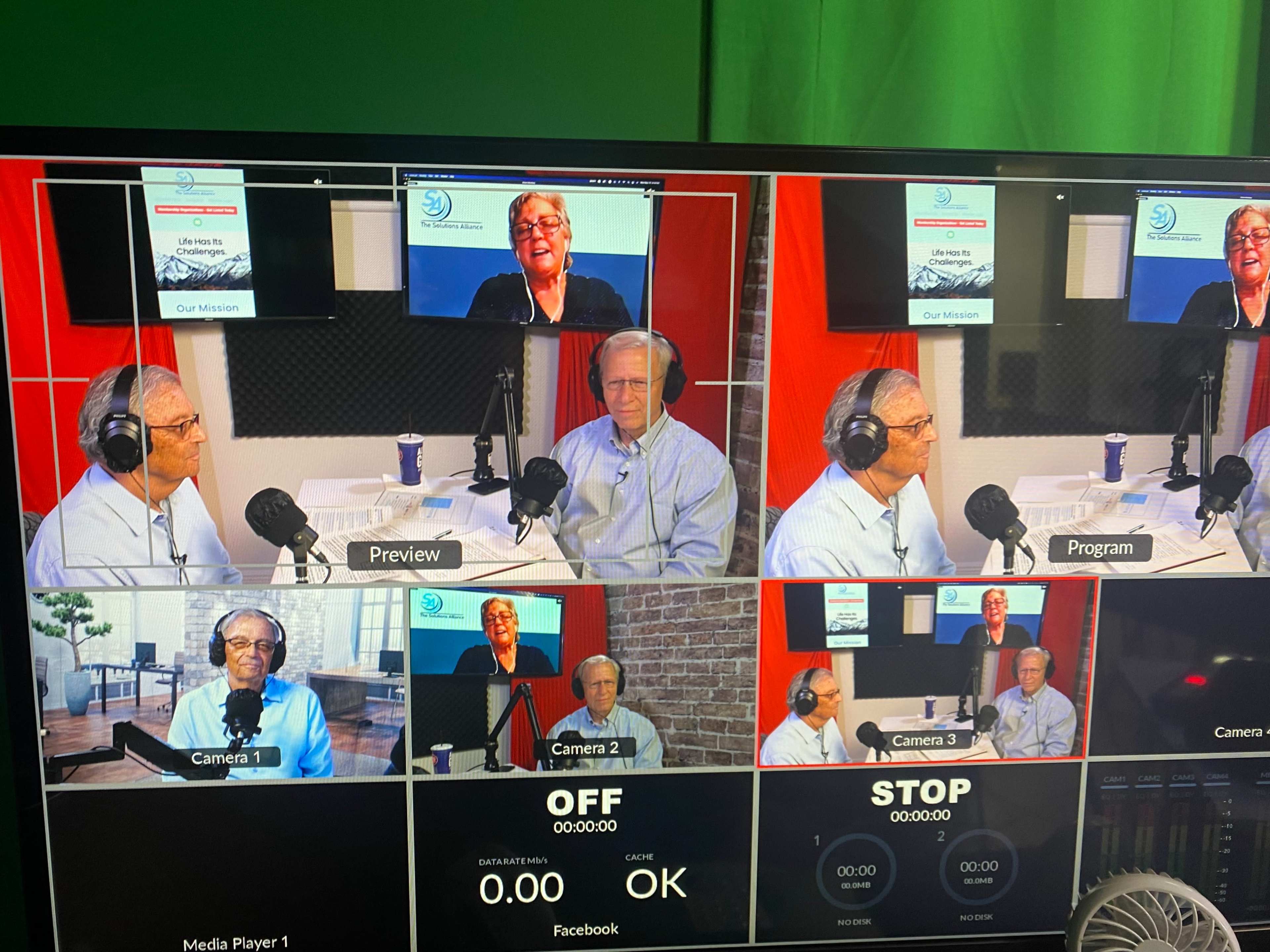 The image shows a video production setup with multiple camera feeds displaying two men engaged in a discussion, alongside a computer screen showing a presentation.