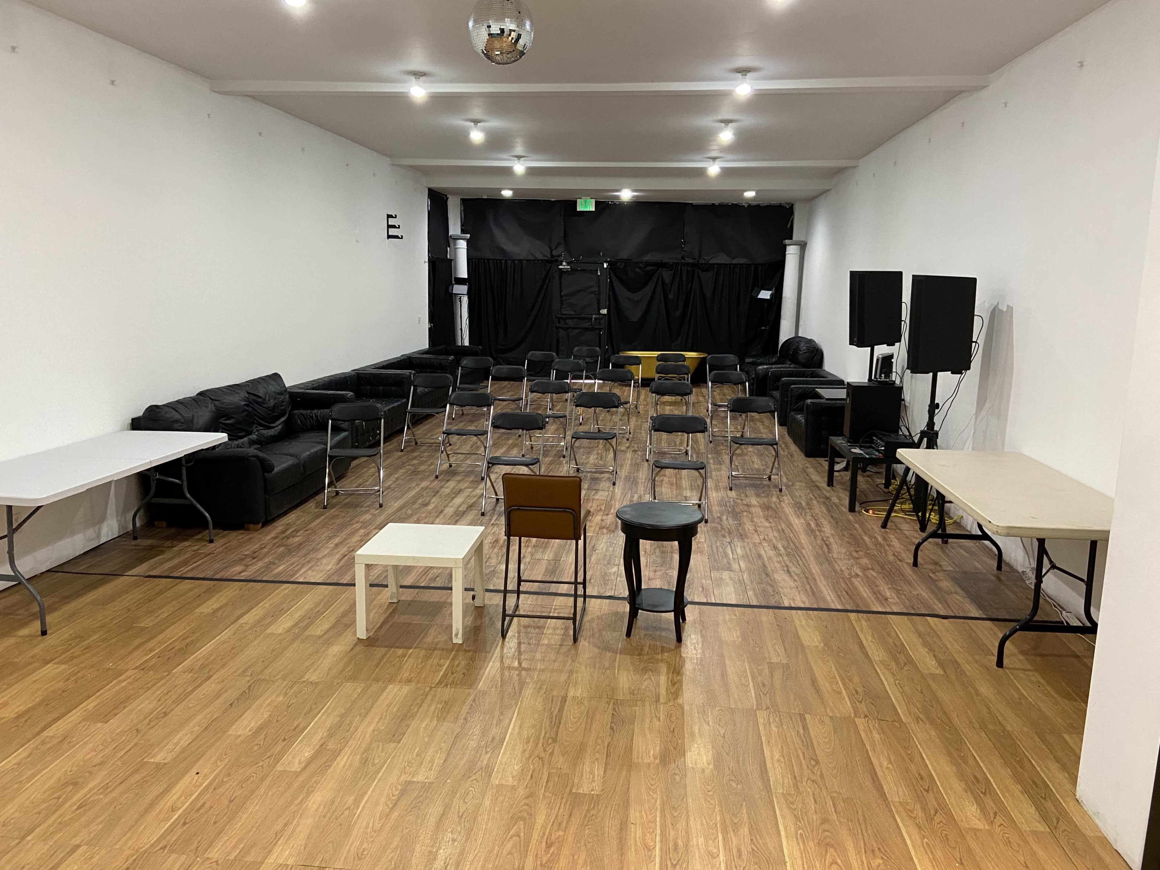 A spacious room with arranged chairs, tables, and black couches against white walls, illuminated by overhead lights.