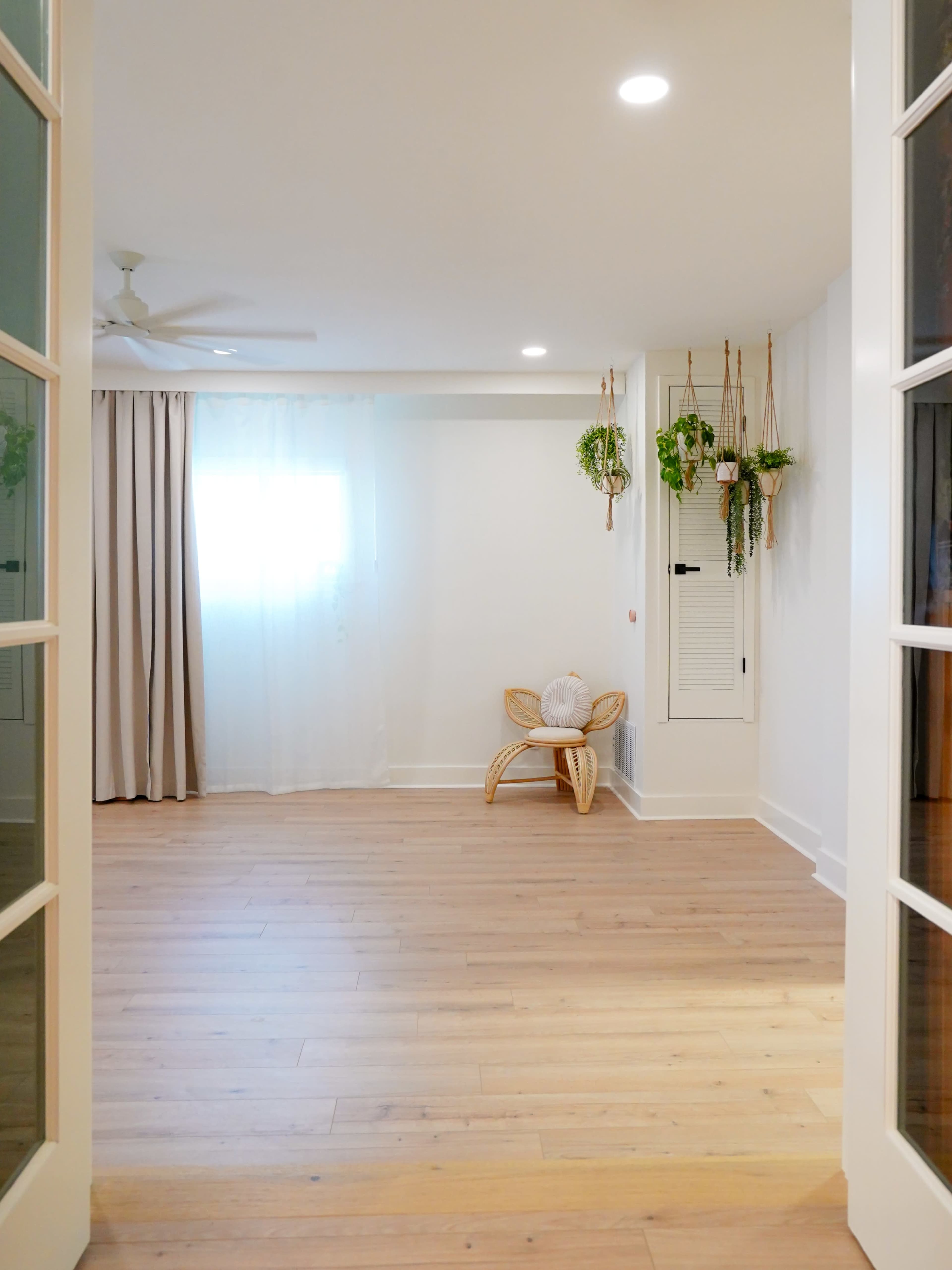 The image depicts a spacious, well-lit room showcasing light-colored wooden flooring, a rattan chair, white curtains, and hanging plants on the walls.