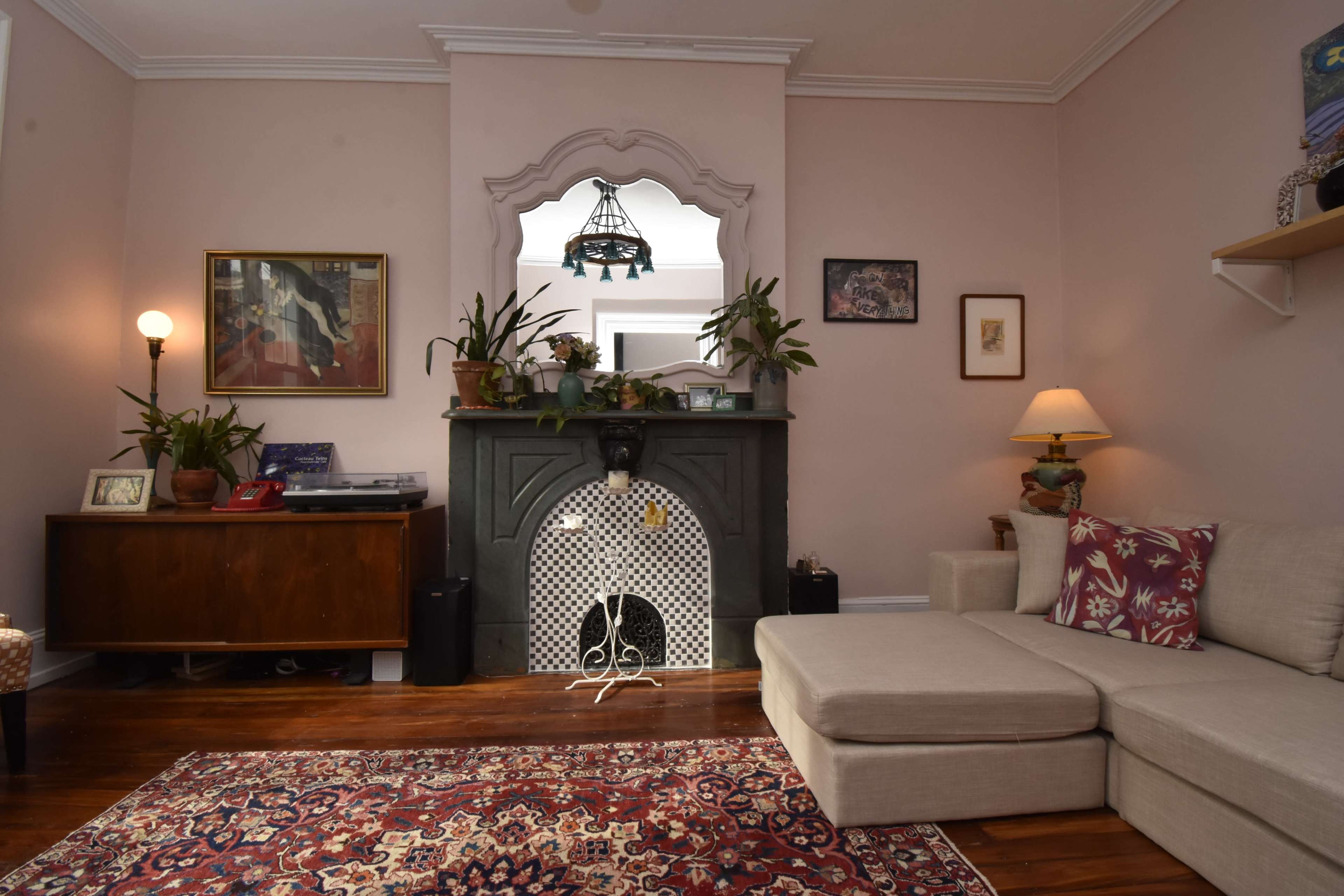 The image shows a cozy living room with a light pink wall, a fireplace centered in the back, and a sectional sofa on the right.