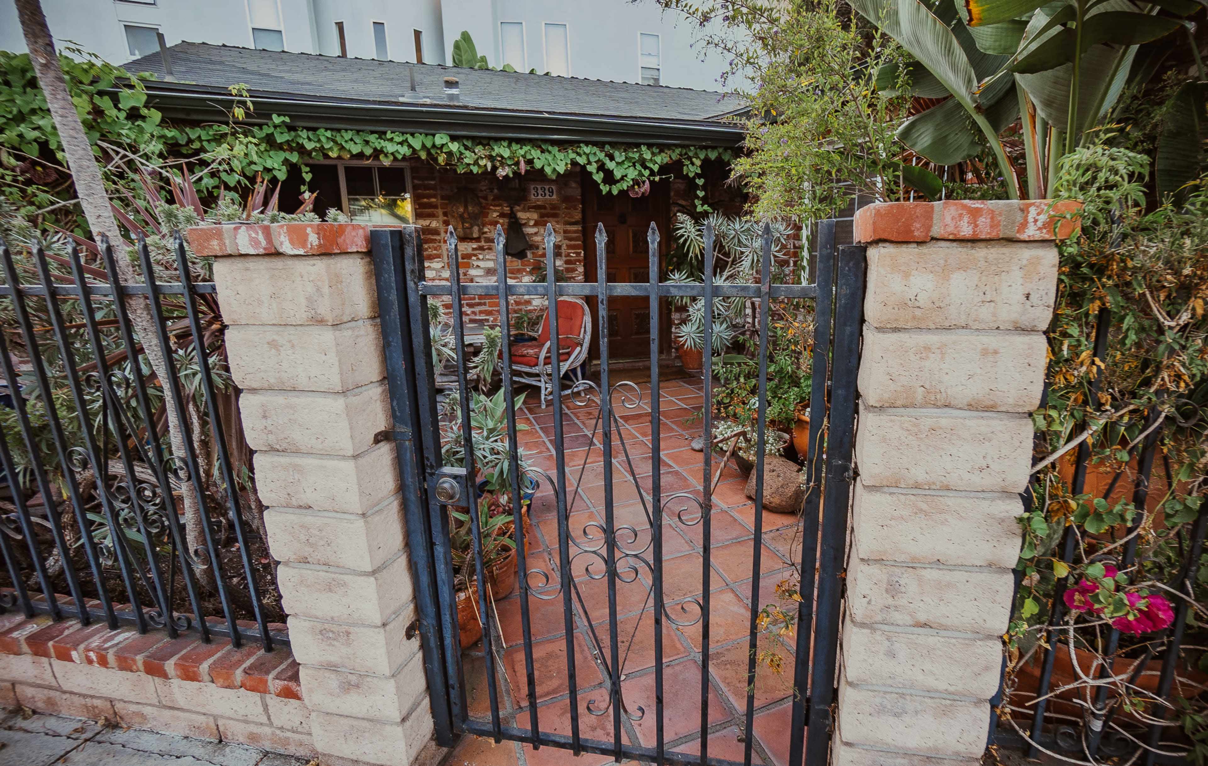A wrought iron gate opens onto a tiled patio surrounded by greenery and potted plants.