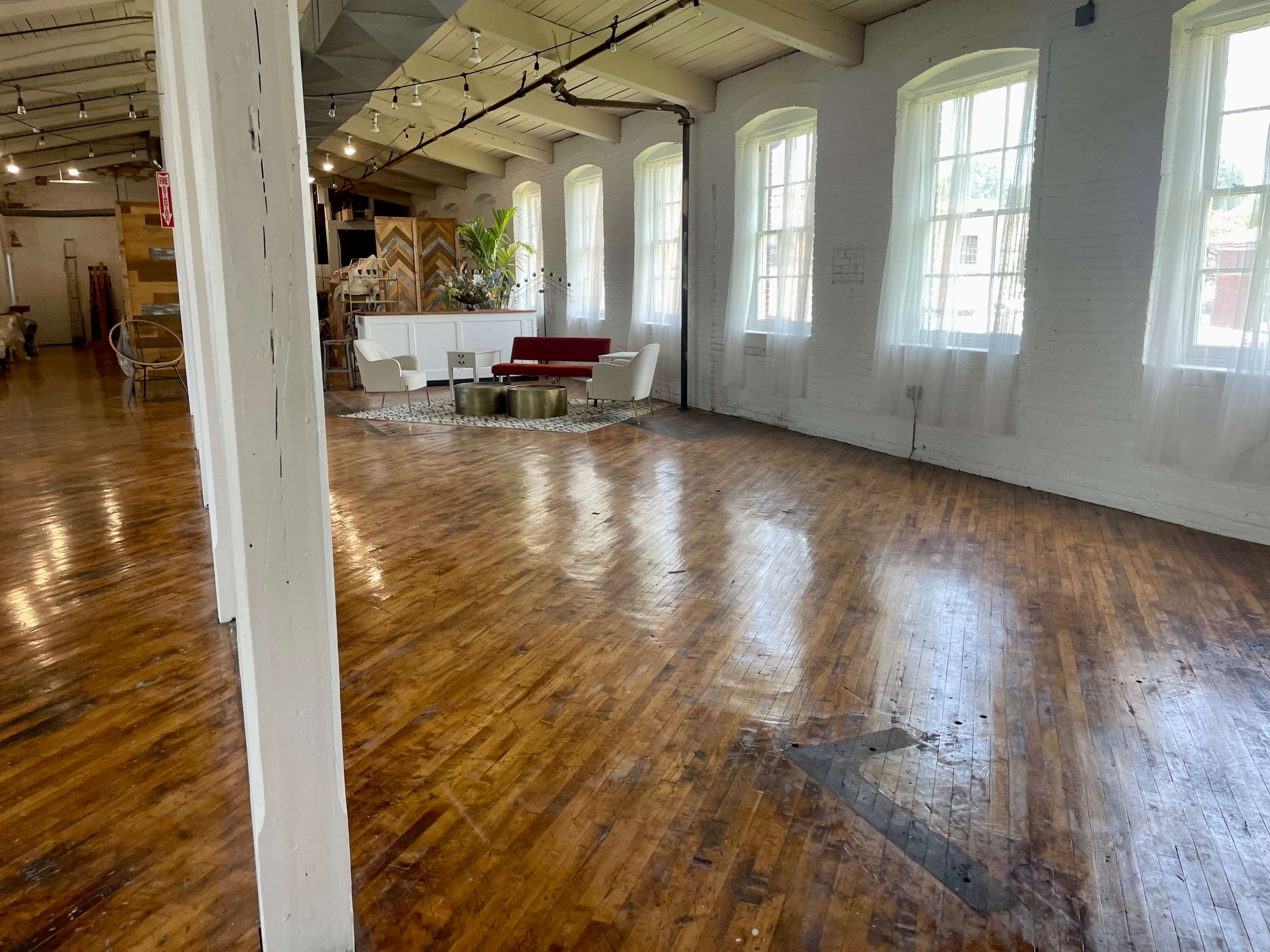 A spacious, well-lit room with wooden flooring, large windows, and a seating area featuring a red couch and white chairs.