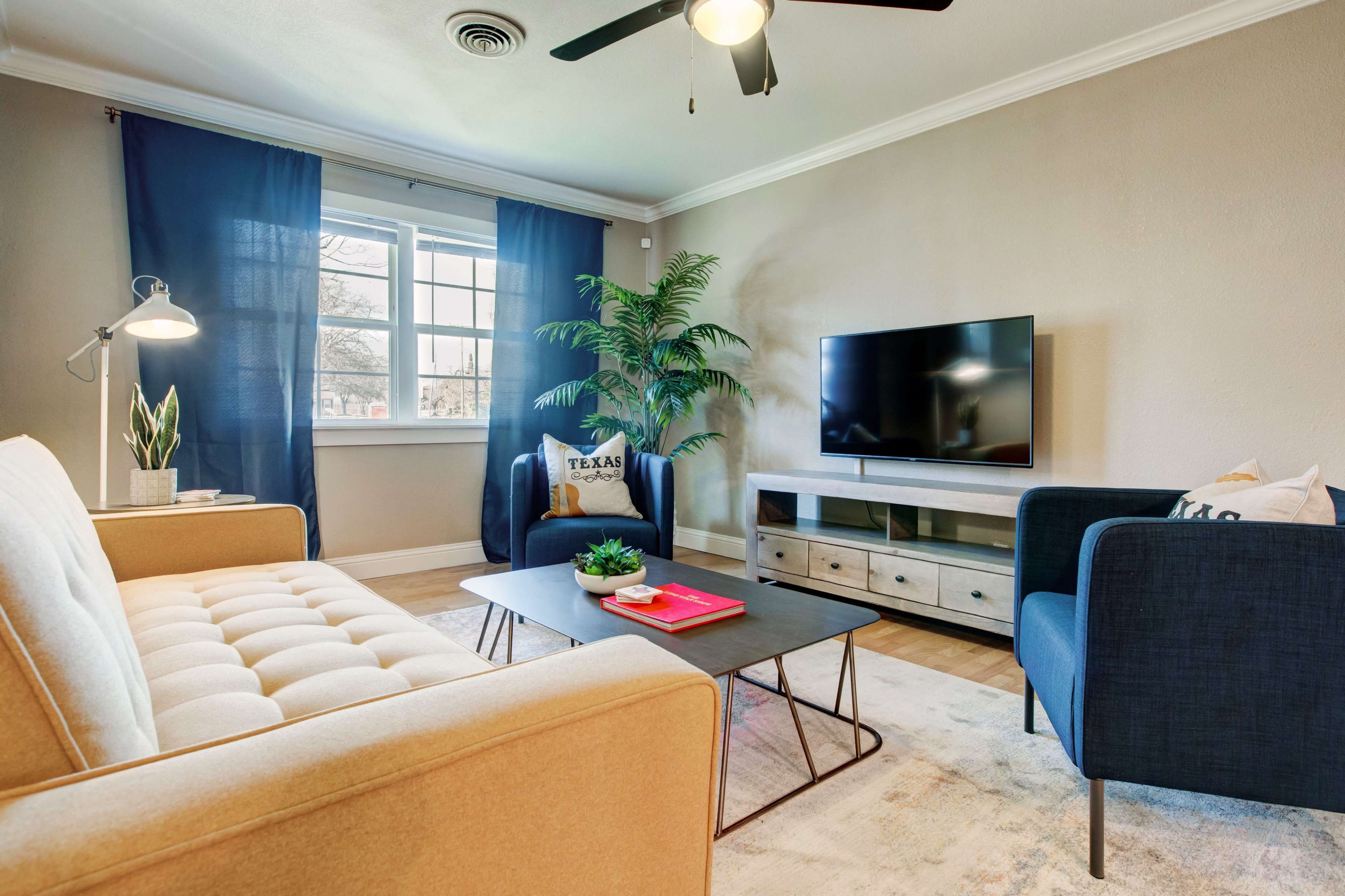 A living room with a beige sofa, a coffee table with books, a flat-screen TV on a cabinet, and potted plants against the backdrop of blue curtains.