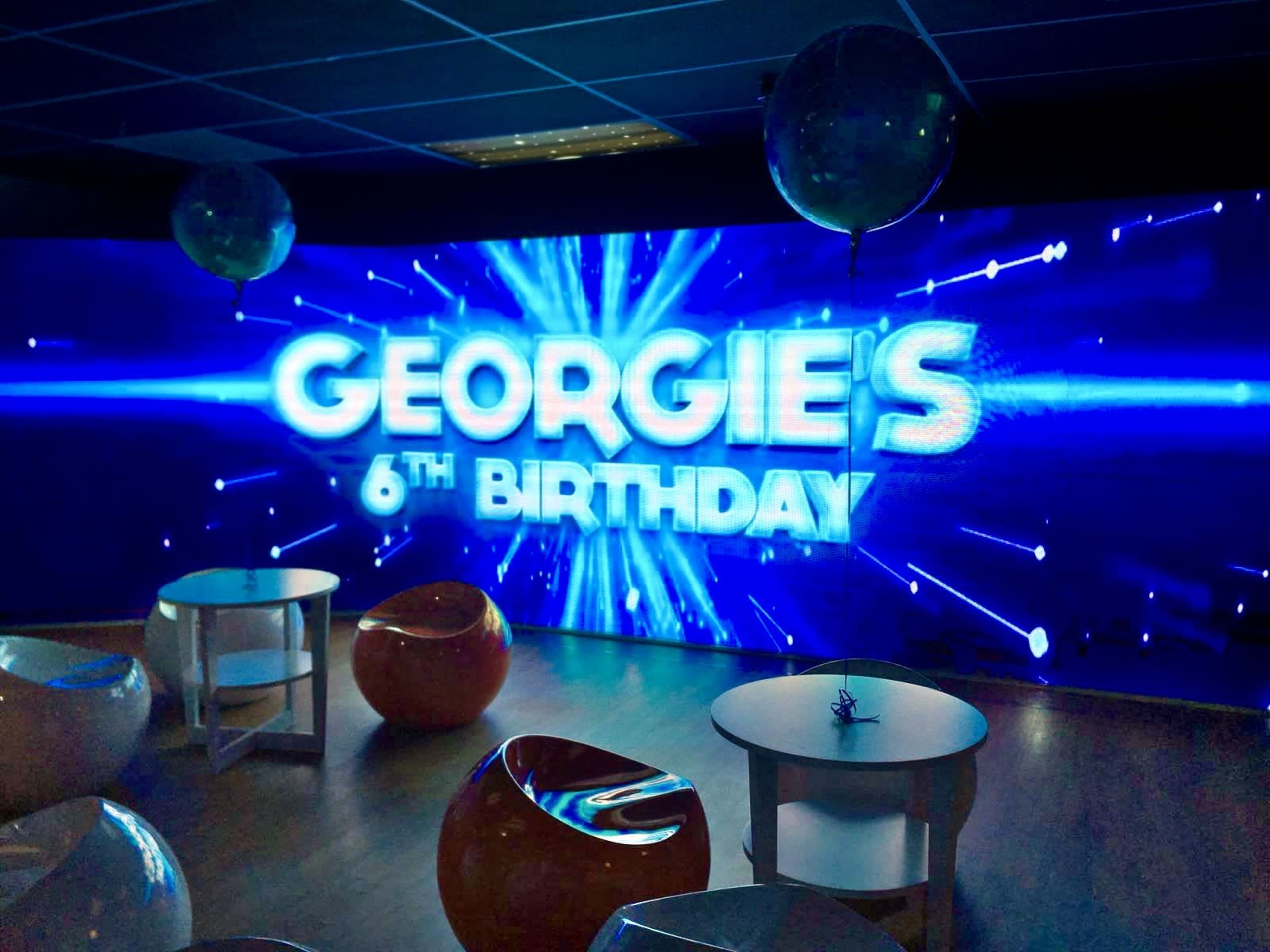 The image features a decorated venue for a birthday party, showcasing a large, illuminated sign that reads "GEORGIE'S 6TH BIRTHDAY" with bright blue lighting effects and a few tables.