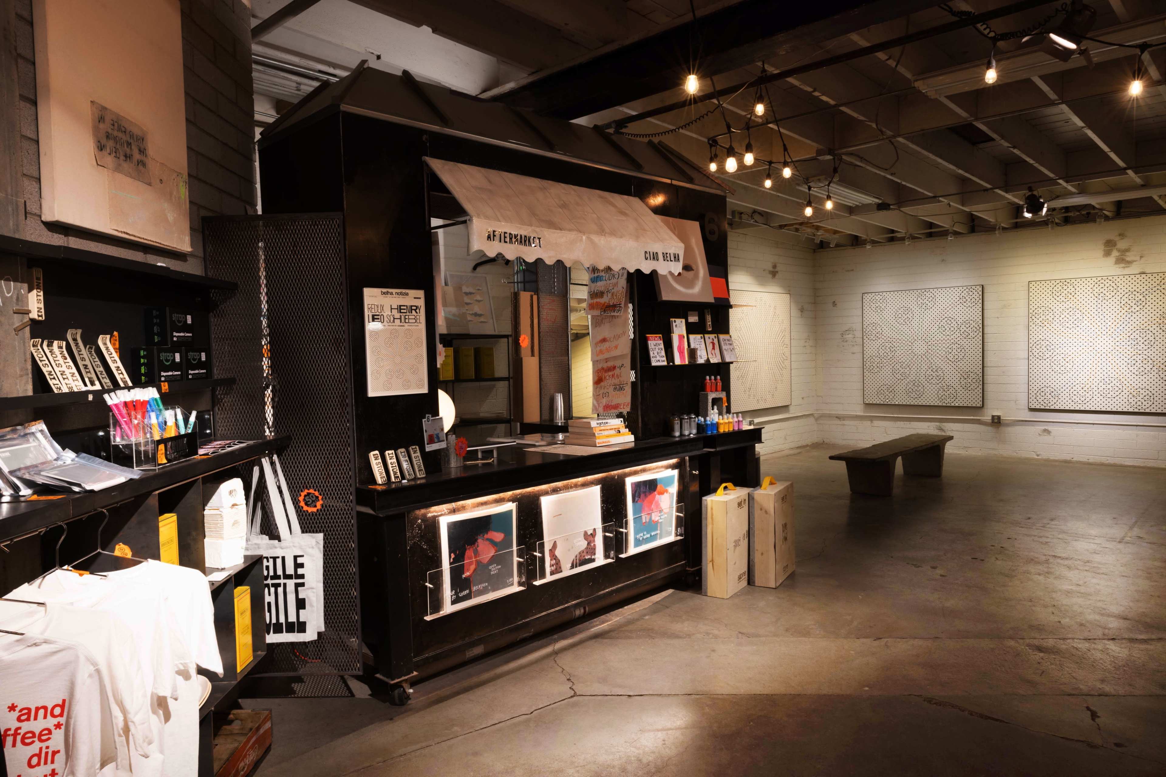 The image shows an art space with a black counter displaying various printed materials and art supplies, under warm overhead lighting.