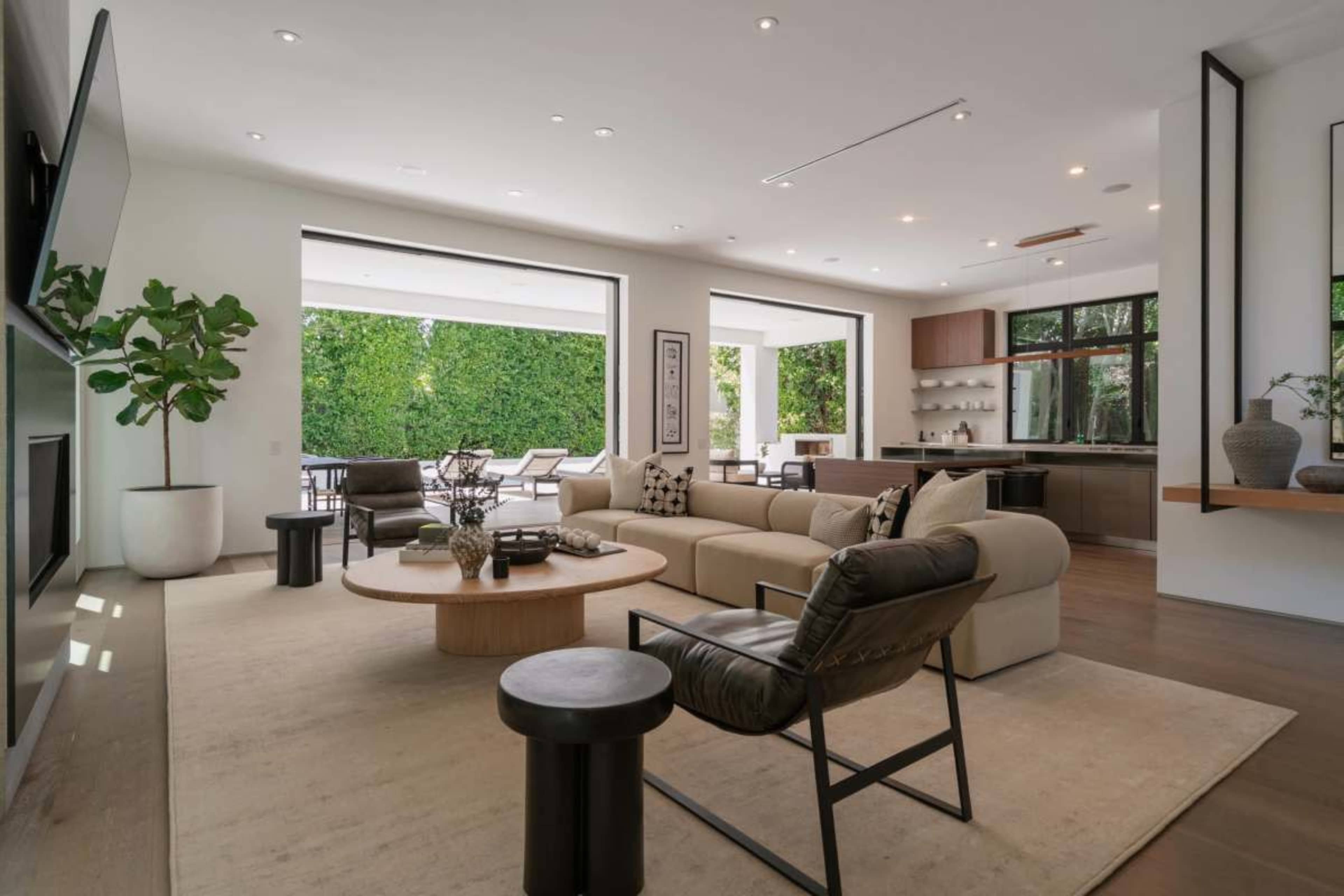 The image shows a modern living room with a light-colored sofa, a round coffee table, and large windows overlooking a green outdoor area.