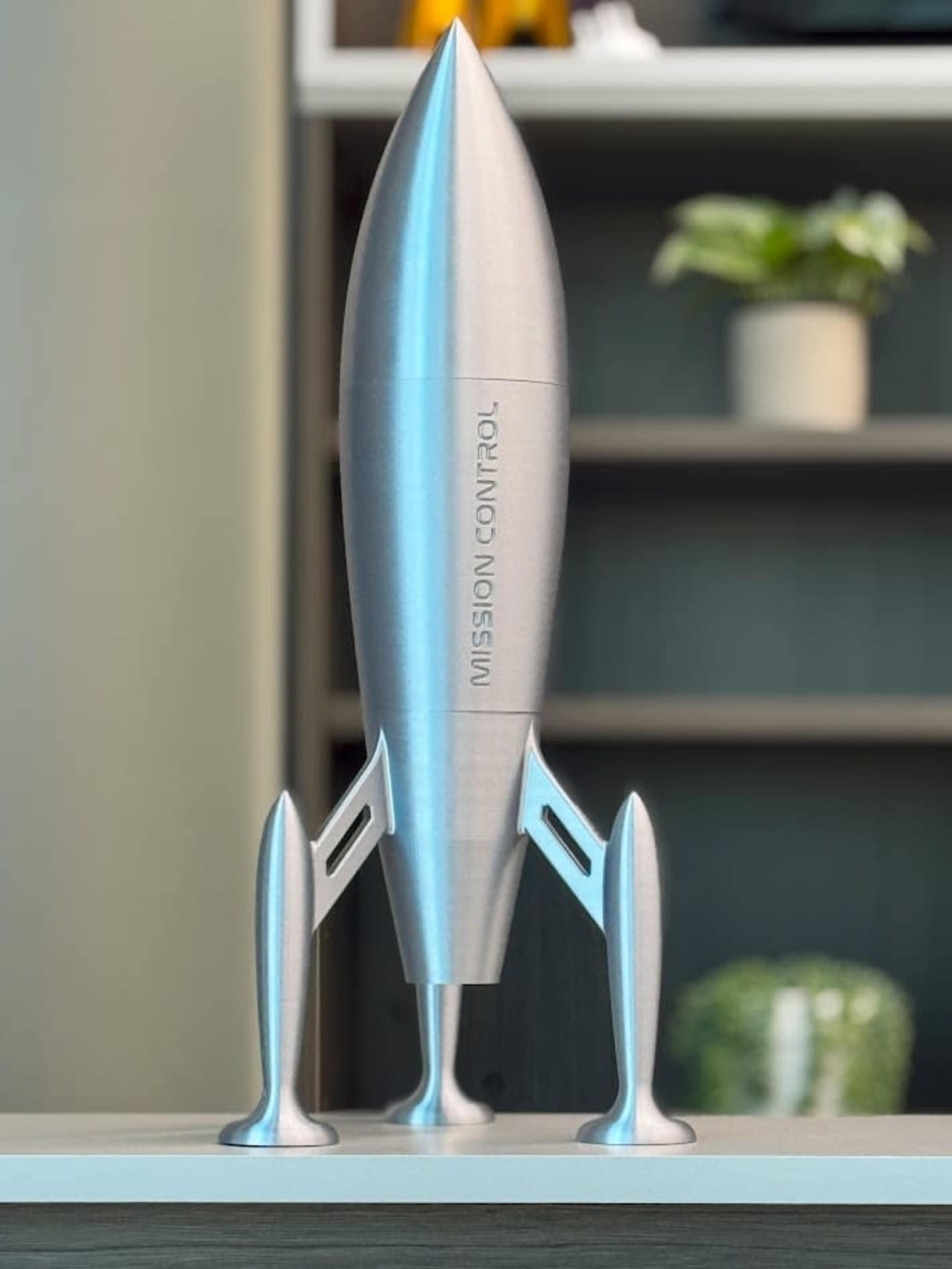 A silver rocket model titled "MISSION CONTROL" is displayed on a shelf with a blurred background featuring a plant and other items.