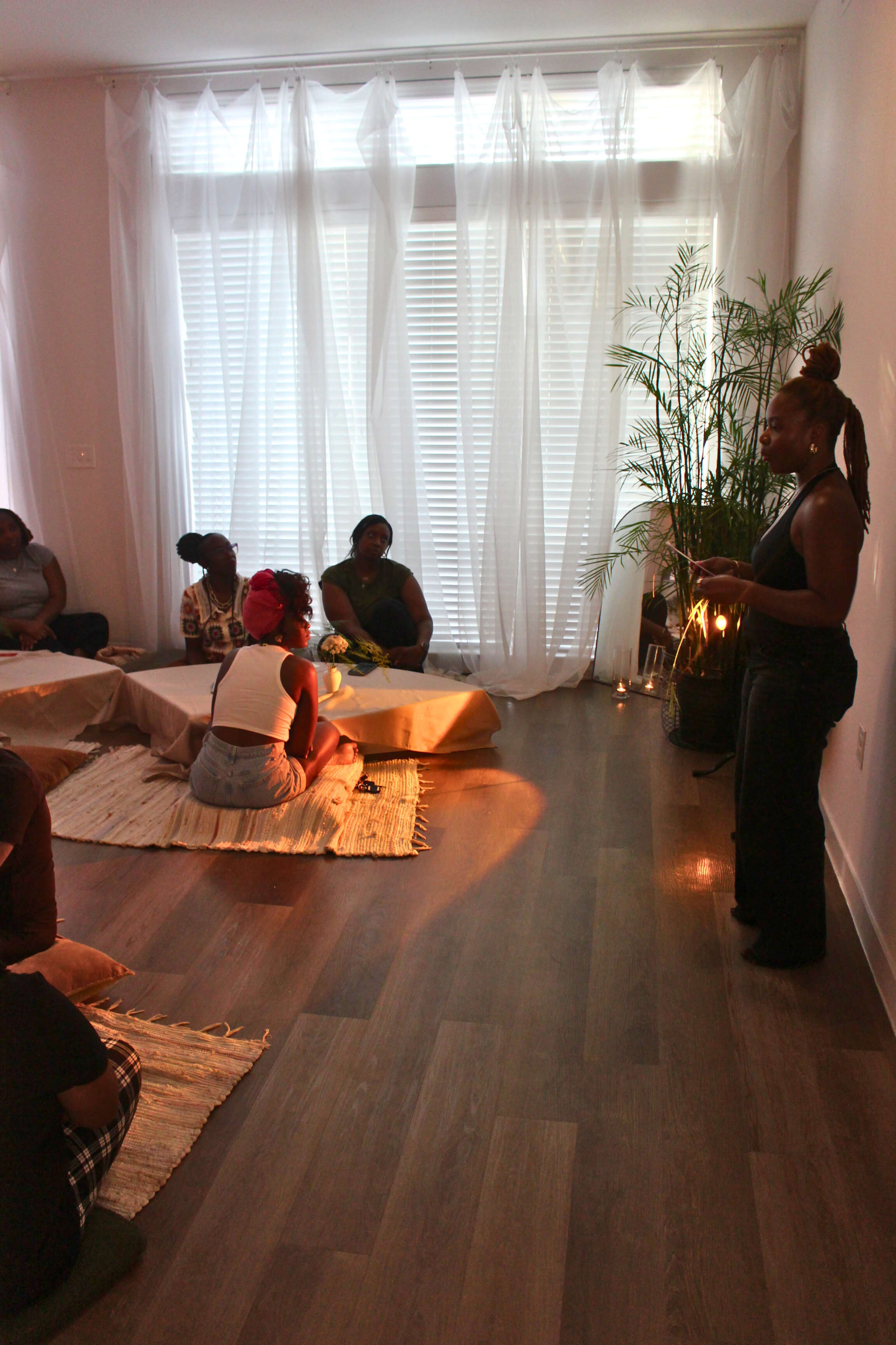 A group of people sits on mats in a bright room with large windows and a plant, while one person stands and speaks to them.