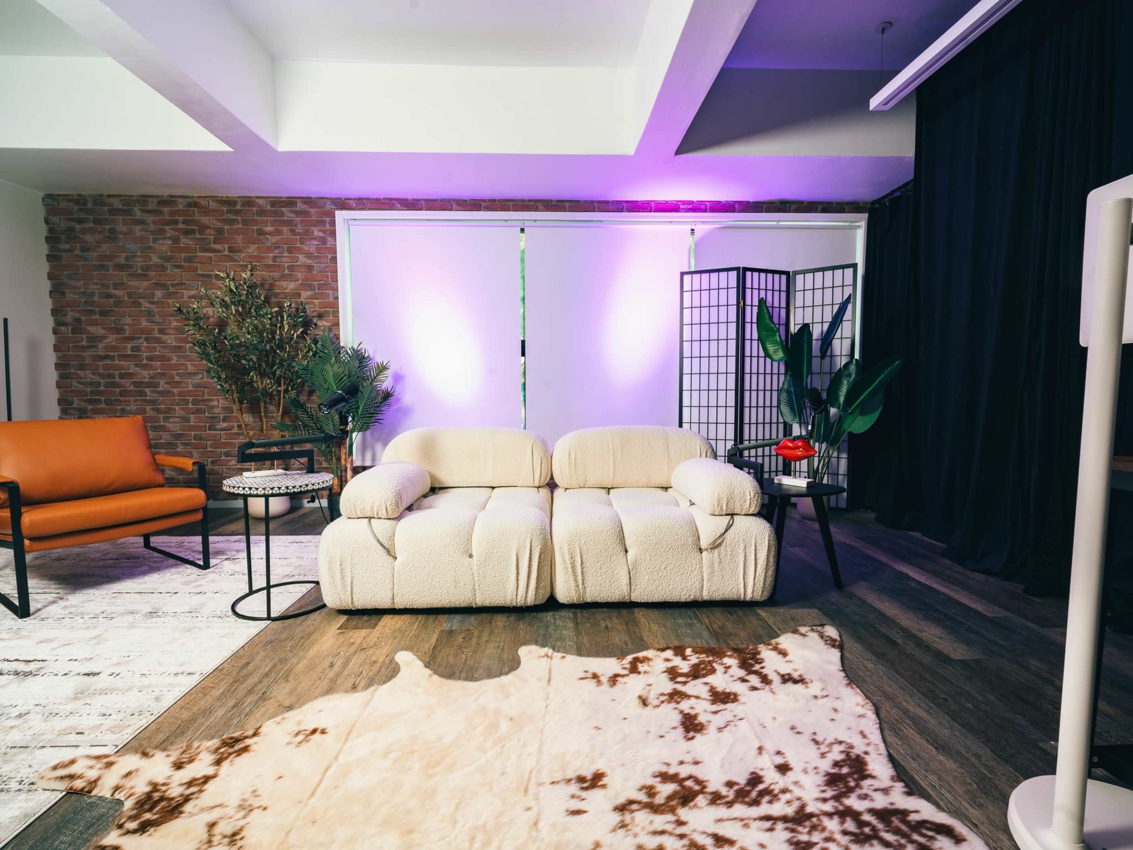 The image shows a modern lounge area featuring a plush cream-colored sofa, an orange armchair, a decorative folding screen, and potted plants, all arranged over a cowhide rug.